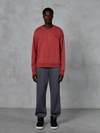 Alpinestars Essentials Ageless Crew, sweatshirt de gola redonda em vermelho tijolo, usada com calças de fato de treino cinzentas e sapatilhas pretas, com um pequeno logótipo preto no peito, streetwear casual inspirado no desporto motorizado