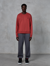 Alpinestars Essentials Ageless Crew, sweatshirt de gola redonda em vermelho tijolo, usada com calças de fato de treino cinzentas e sapatilhas pretas, com um pequeno logótipo preto no peito, streetwear casual inspirado no desporto motorizado