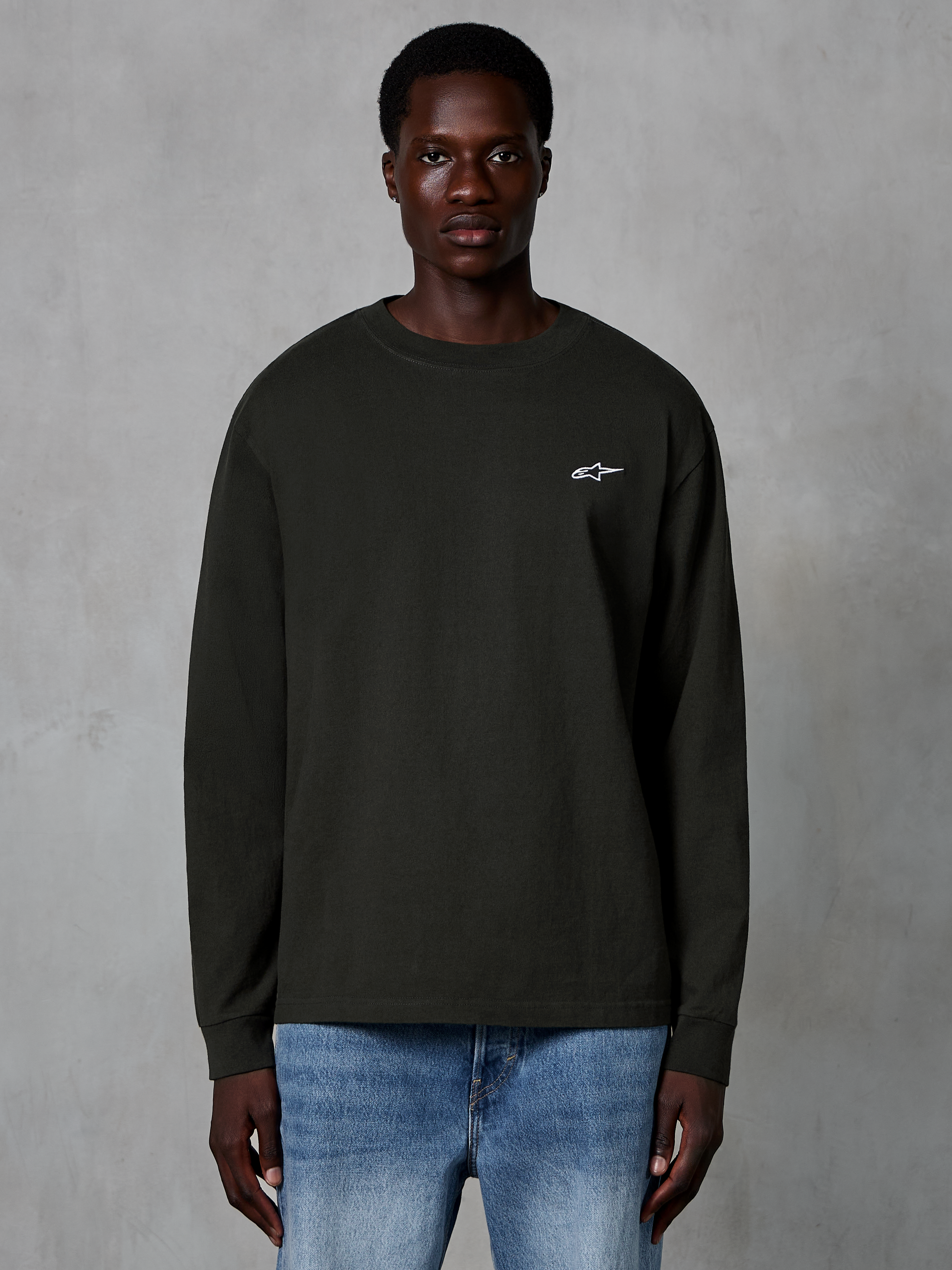 Alpinestars Essentials Ageless LS Tee, t-shirt de manga comprida, Off Black, com um pequeno logótipo Alpinestars branco no peito, vestida por um modelo com calças de ganga azuis, streetwear casual de algodão orgânico