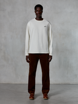 Alpinestars Essentials Ageless LS Tee, t-shirt de manga comprida, Off-White, vestida por um modelo com calças de bombazina castanhas e botas de pele castanhas, com um pequeno logótipo preto no peito, corte contemporâneo e relaxado, estilo casual lifestyle streetwear