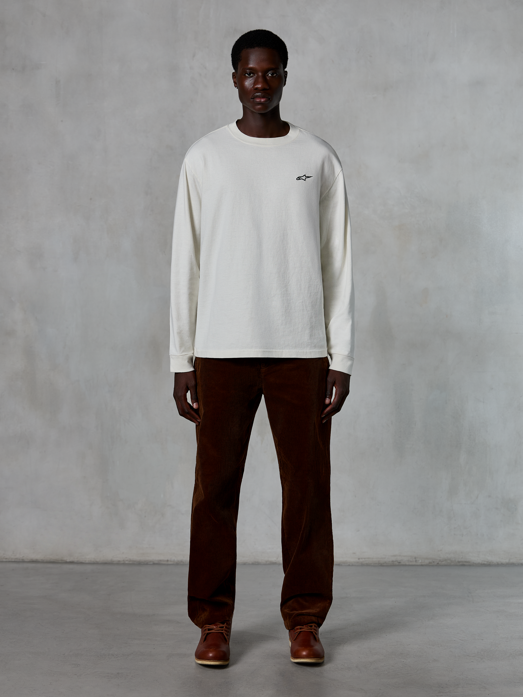 Alpinestars Essentials Ageless LS Tee, t-shirt de manga comprida, Off-White, vestida por um modelo com calças de bombazina castanhas e botas de pele castanhas, com um pequeno logótipo preto no peito, corte contemporâneo e relaxado, estilo casual lifestyle streetwear