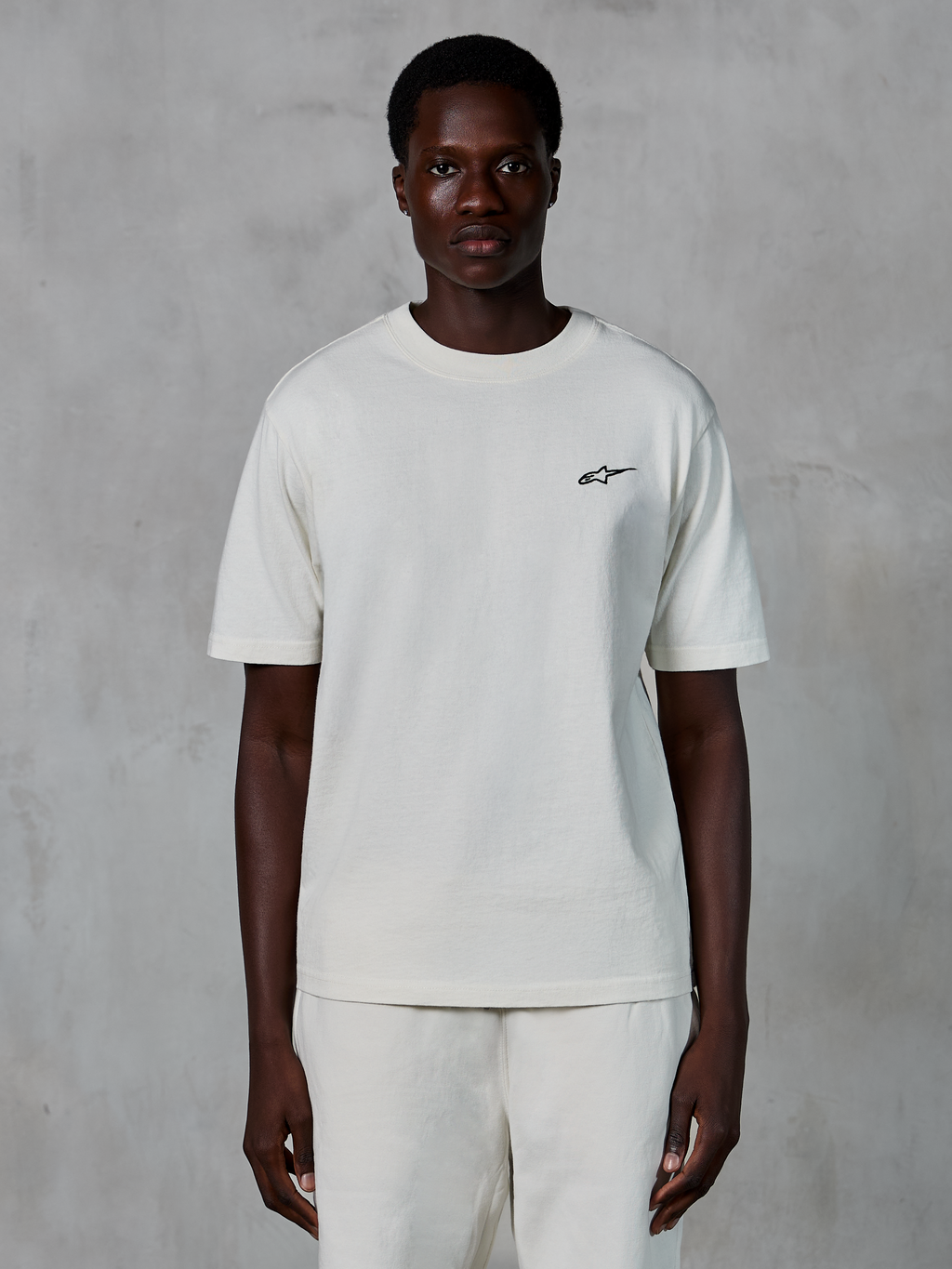 Alpinestars Essentials Ageless SS Tee, t-shirt de manga curta, Off White, usada por um modelo masculino, com um pequeno logótipo Alpinestars preto no peito, corte relaxado, jersey de algodão orgânico, streetwear casual de desportos motorizados