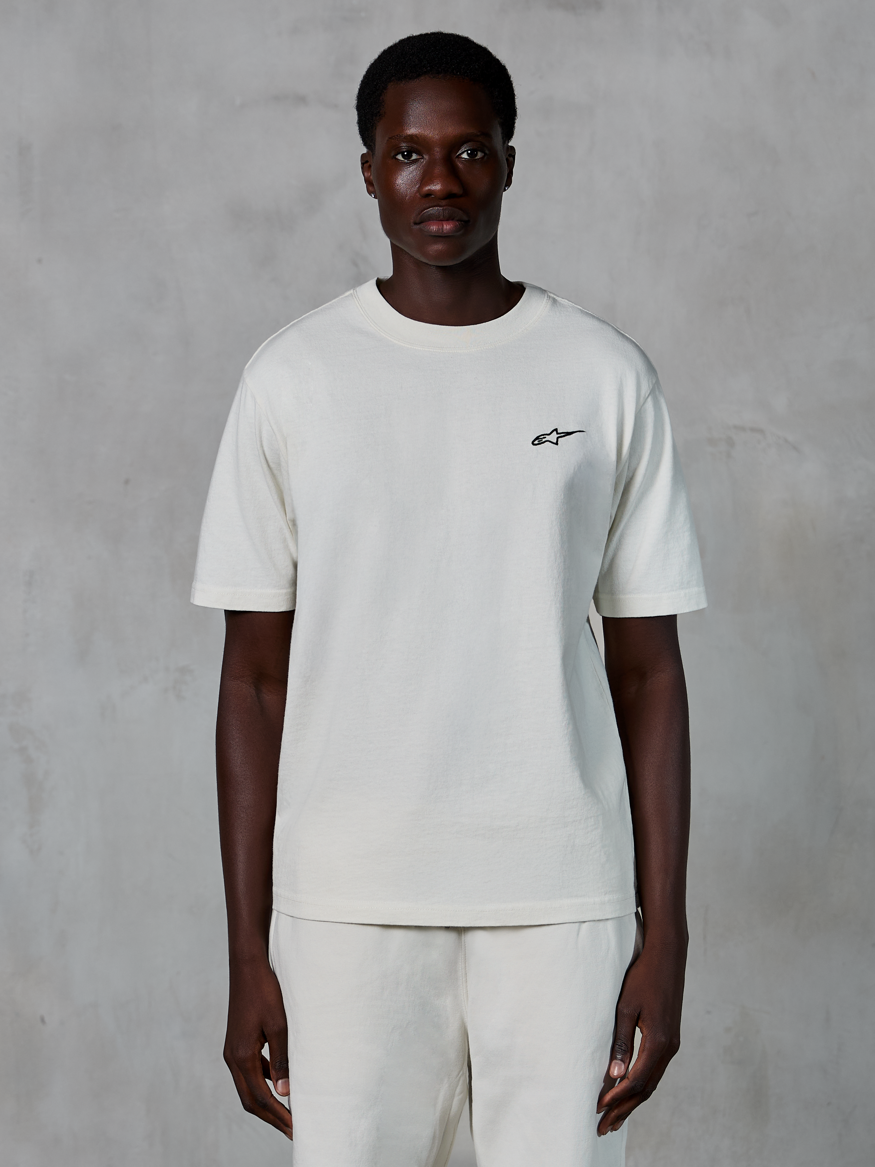 Alpinestars Essentials Ageless SS Tee, t-shirt de manga curta, Off White, usada por um modelo masculino, com um pequeno logótipo Alpinestars preto no peito, corte relaxado, jersey de algodão orgânico, streetwear casual de desportos motorizados
