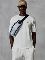 Alpinestars Essentials Ageless SS Tee, t-shirt de manga curta, Off White, usada com calções a condizer e um boné bege, com um pequeno logótipo preto no peito e uma bolsa de tiracolo cinzenta, streetwear casual inspirado no desporto motorizado