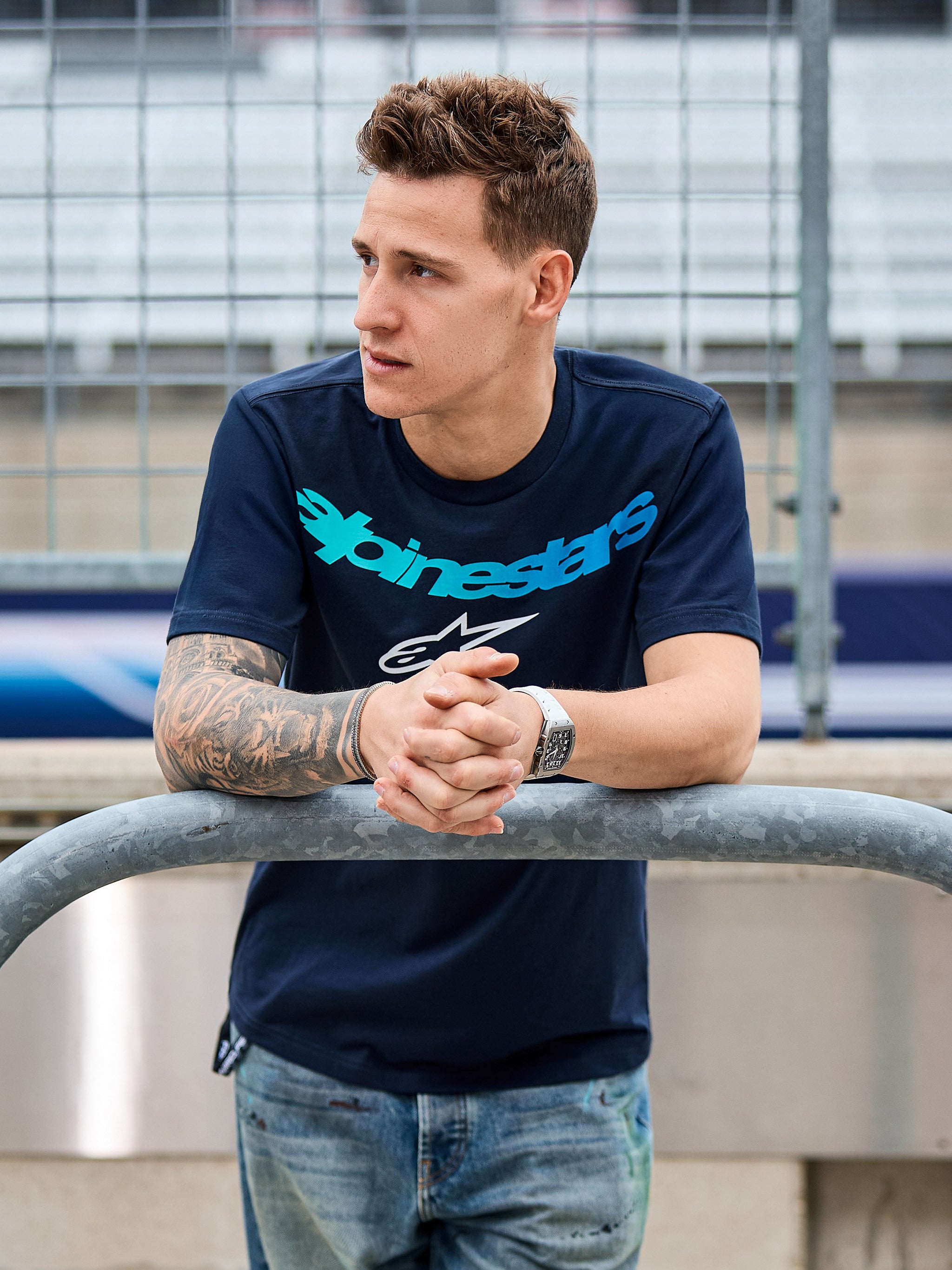 T-shirt Alpinestars Collared CSF, camisola de manga curta, azul-marinho, texto Alpinestars em azul-petróleo e logótipo de estrela branco, usada por um homem encostado a um corrimão metálico num circuito, casual motorsport streetwear