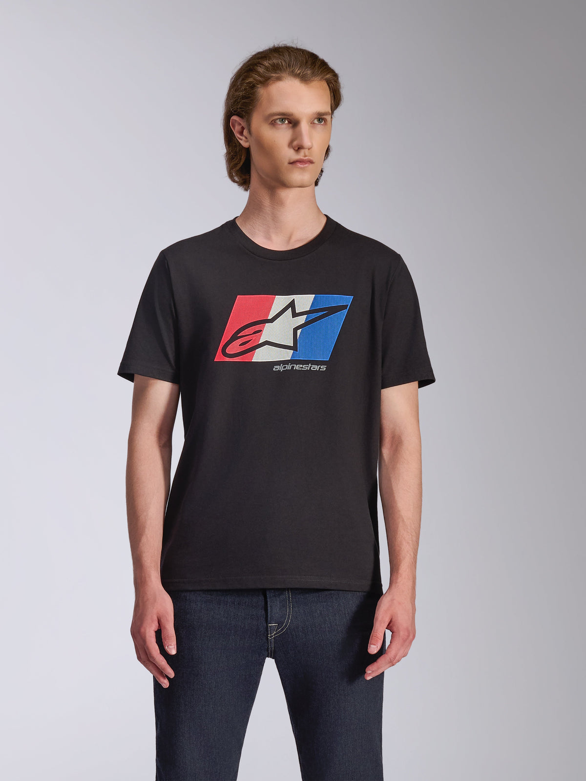 Alpinestars Global CSF Tee, T-shirt de manga curta, preta, gráfico do logótipo Ageless em vermelho, branco e azul, vestida por um modelo masculino com calças de ganga escuras, streetwear casual de desportos motorizados