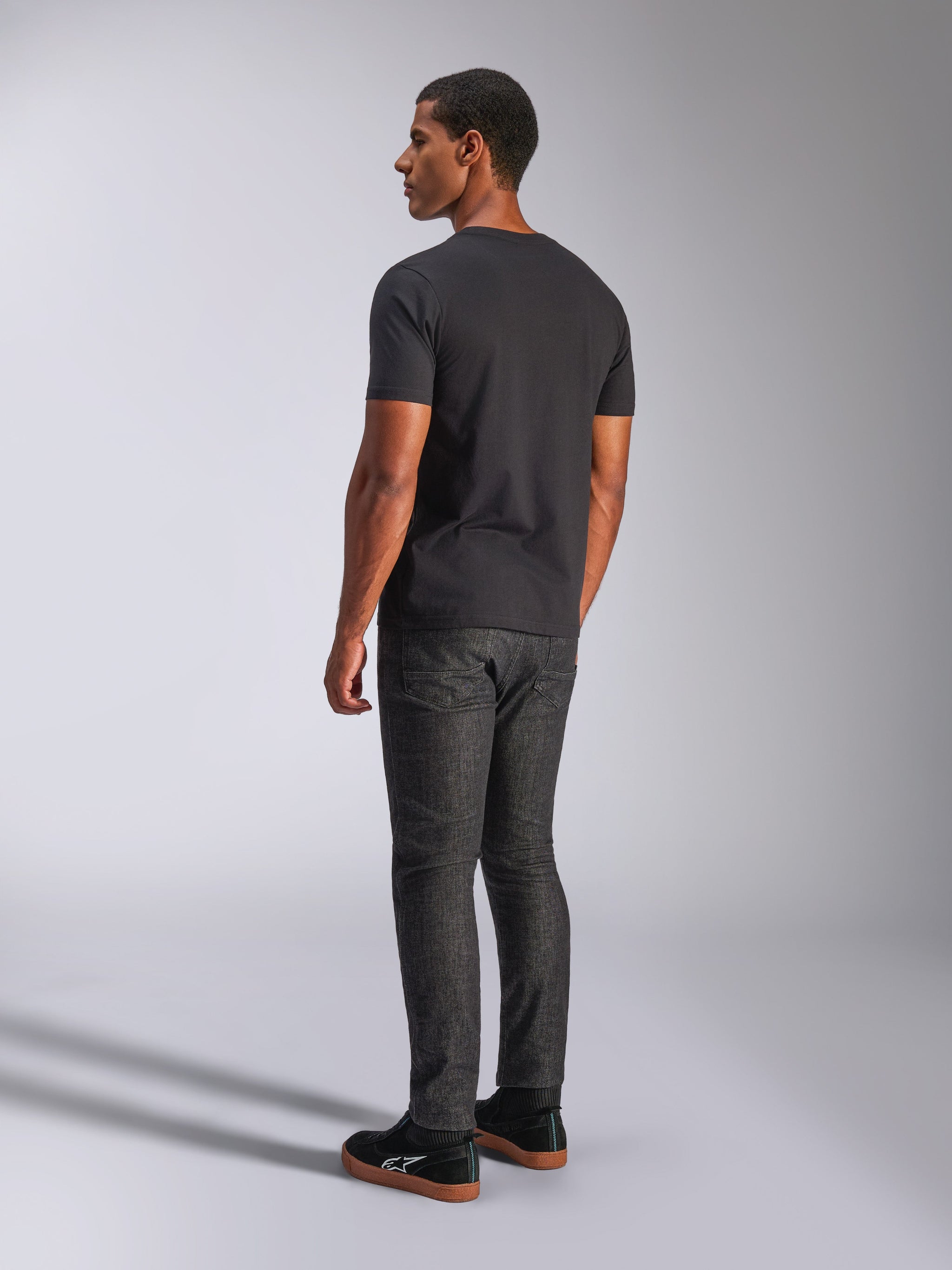 Alpinestars Tech Boot CSF Tee, t-shirt de manga curta, preta, vista traseira no modelo, usada com calças de ganga cinzento escuro e sapatilhas pretas, estilo casual motorsport streetwear
