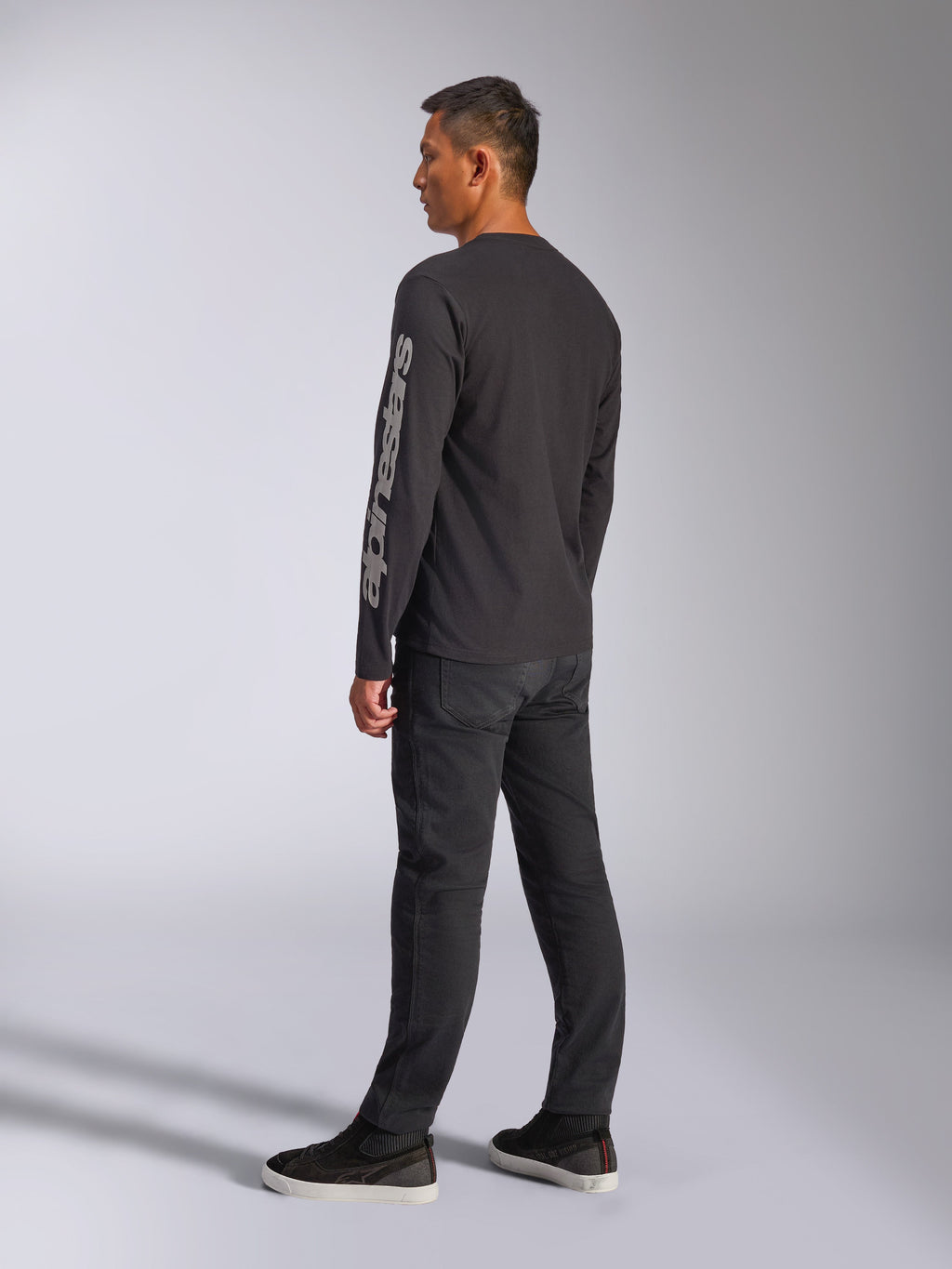 T-shirt de manga comprida Alpinestars Trackside CSF, preta, com um grande logótipo Alpinestars cinzento na manga esquerda, usada por um modelo com calças pretas e sapatilhas casuais, estilo streetwear casual inspirado no desporto motorizado.