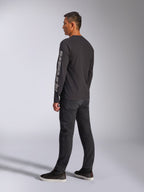 T-shirt de manga comprida Alpinestars Trackside CSF, preta, com um grande logótipo Alpinestars cinzento na manga esquerda, usada por um modelo com calças pretas e sapatilhas casuais, estilo streetwear casual inspirado no desporto motorizado.