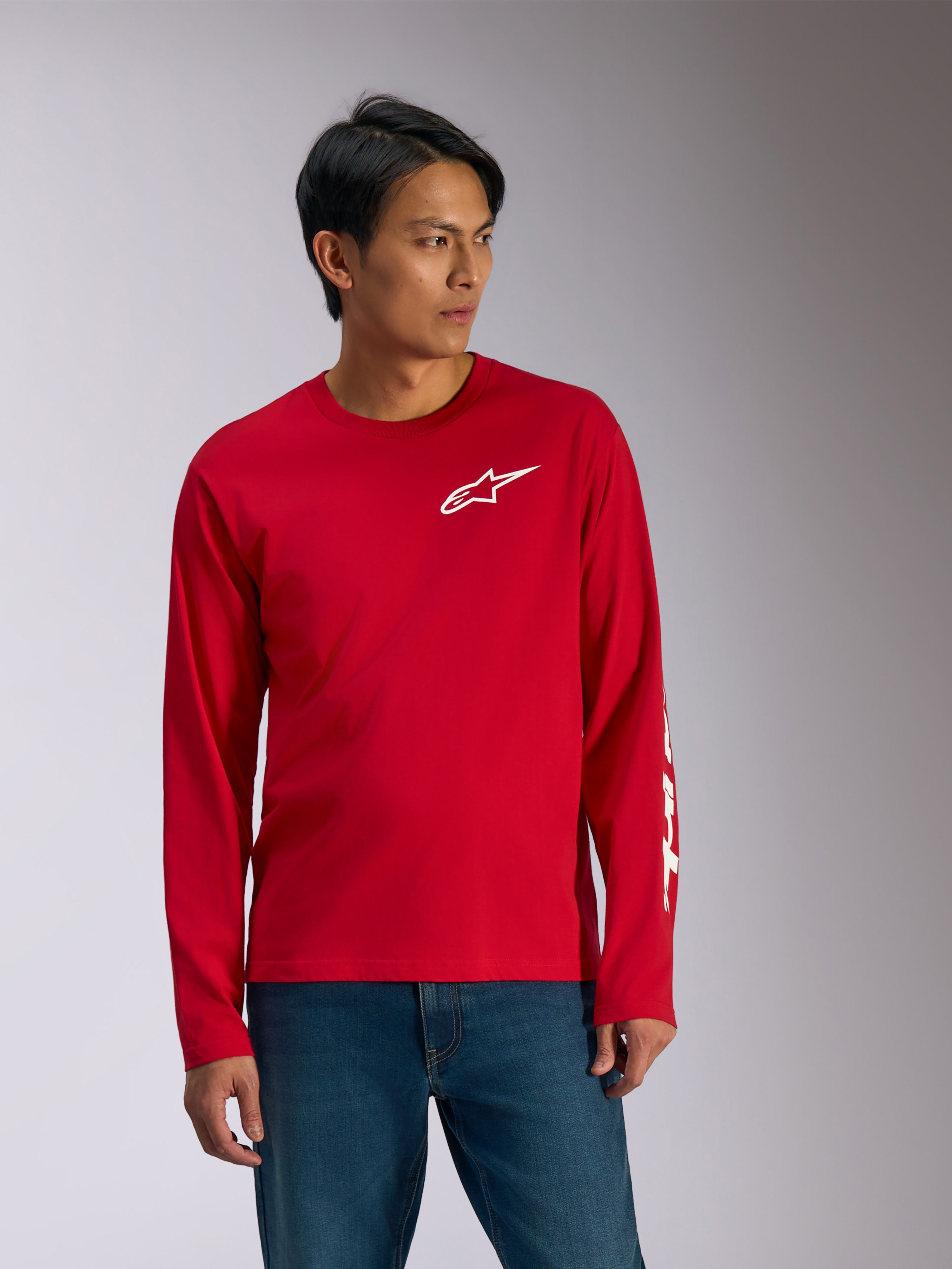 Alpinestars Trackside CSF Tee, t-shirt de manga comprida, vermelha com grafismos brancos, logótipo de estrela da Alpinestars branco no peito e texto branco na manga, usada com calças de ganga azuis, vestuário desportivo casual inspirado no desporto motorizado