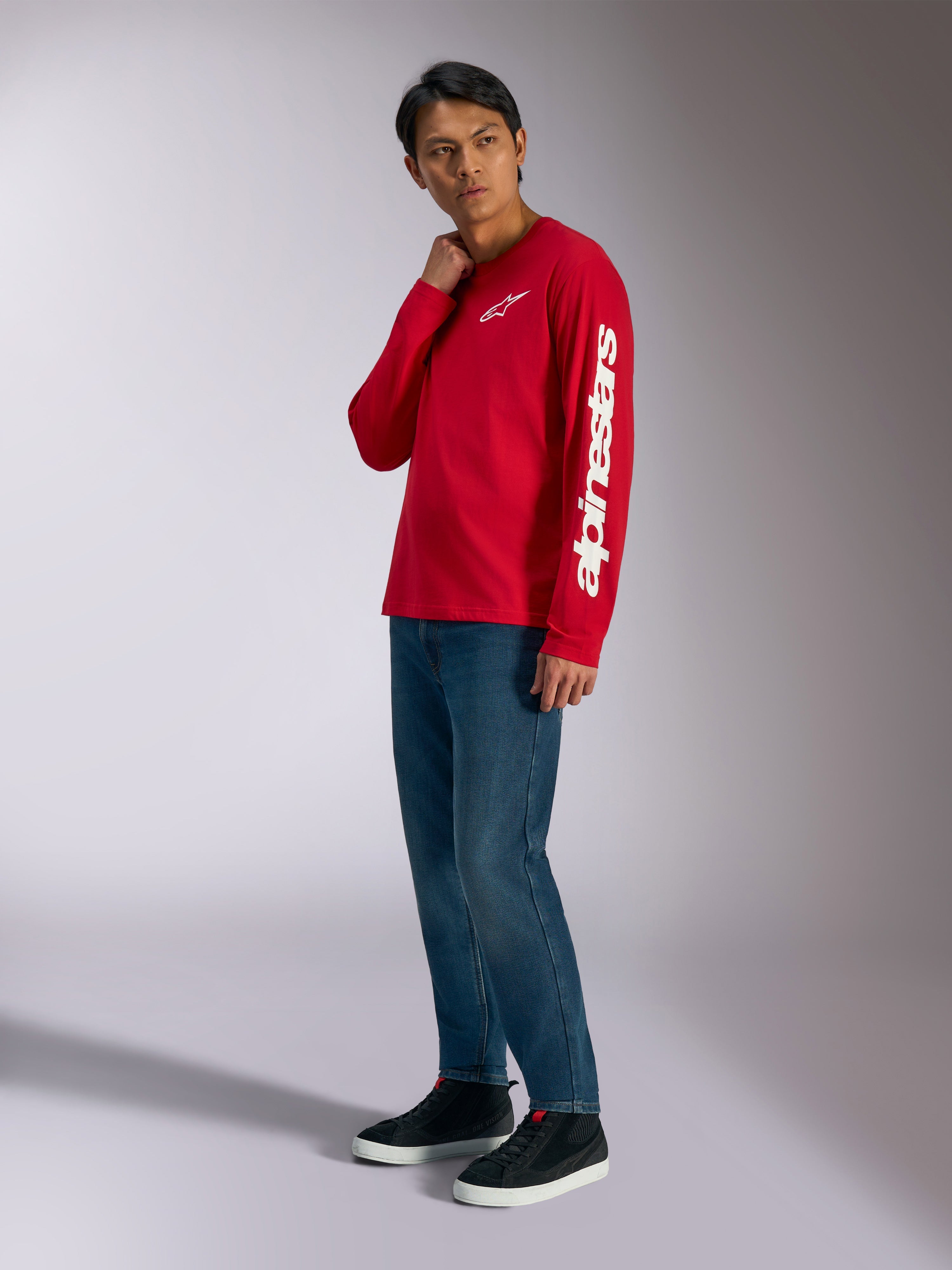 T-shirt de manga comprida Alpinestars Trackside CSF, vermelha com logótipos Alpinestars brancos no peito e na manga, usada por modelo com calças de ganga azuis e sapatilhas pretas, streetwear casual inspirado no desporto motorizado