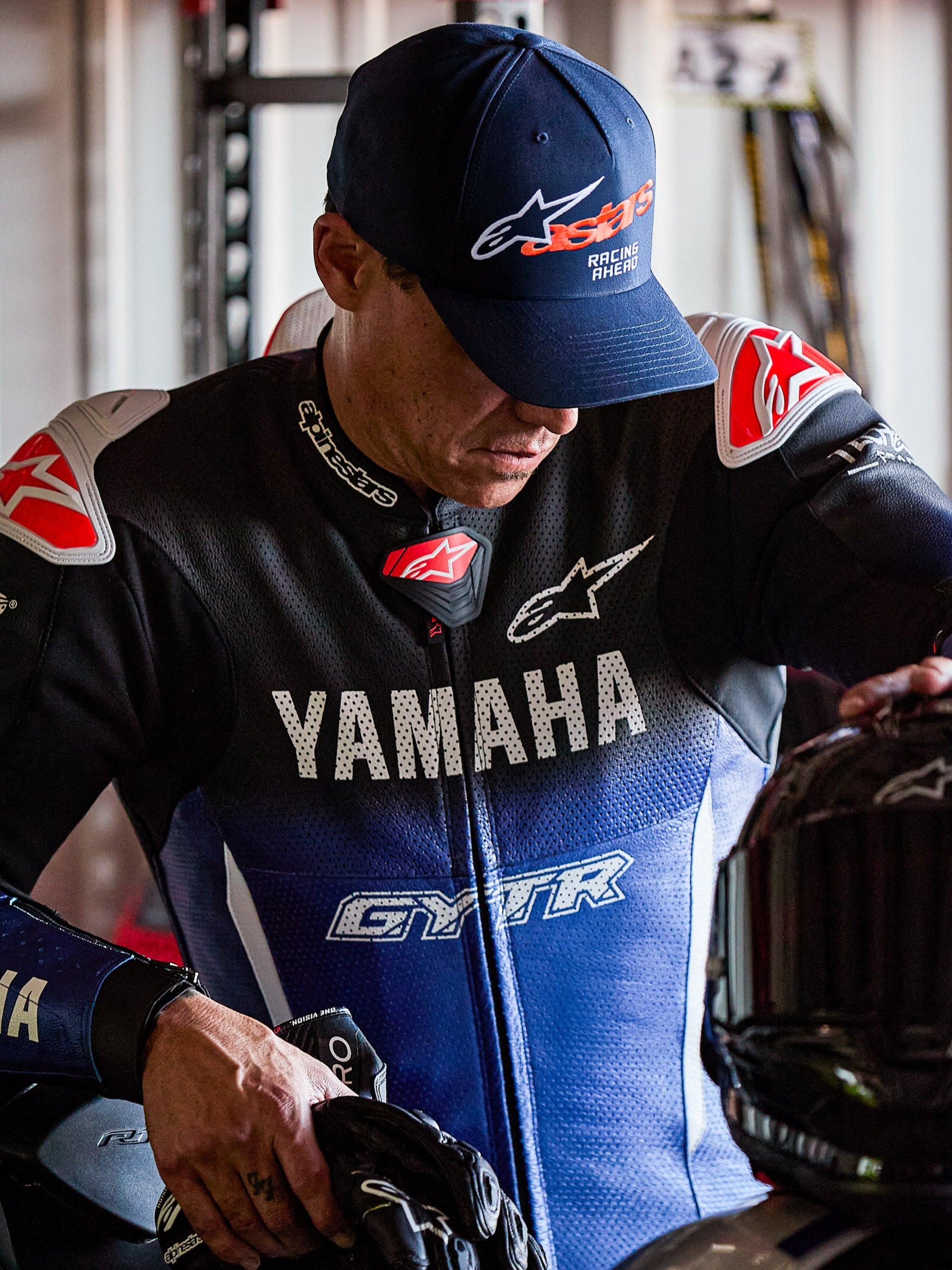 Boné Alpinestars Entitled, azul-marinho com logótipo Astars laranja e texto 'Racing Ahead' em branco, usado por um piloto num fato de corrida Yamaha azul e preto, boné de basebol de aba curva com coroa estruturada.