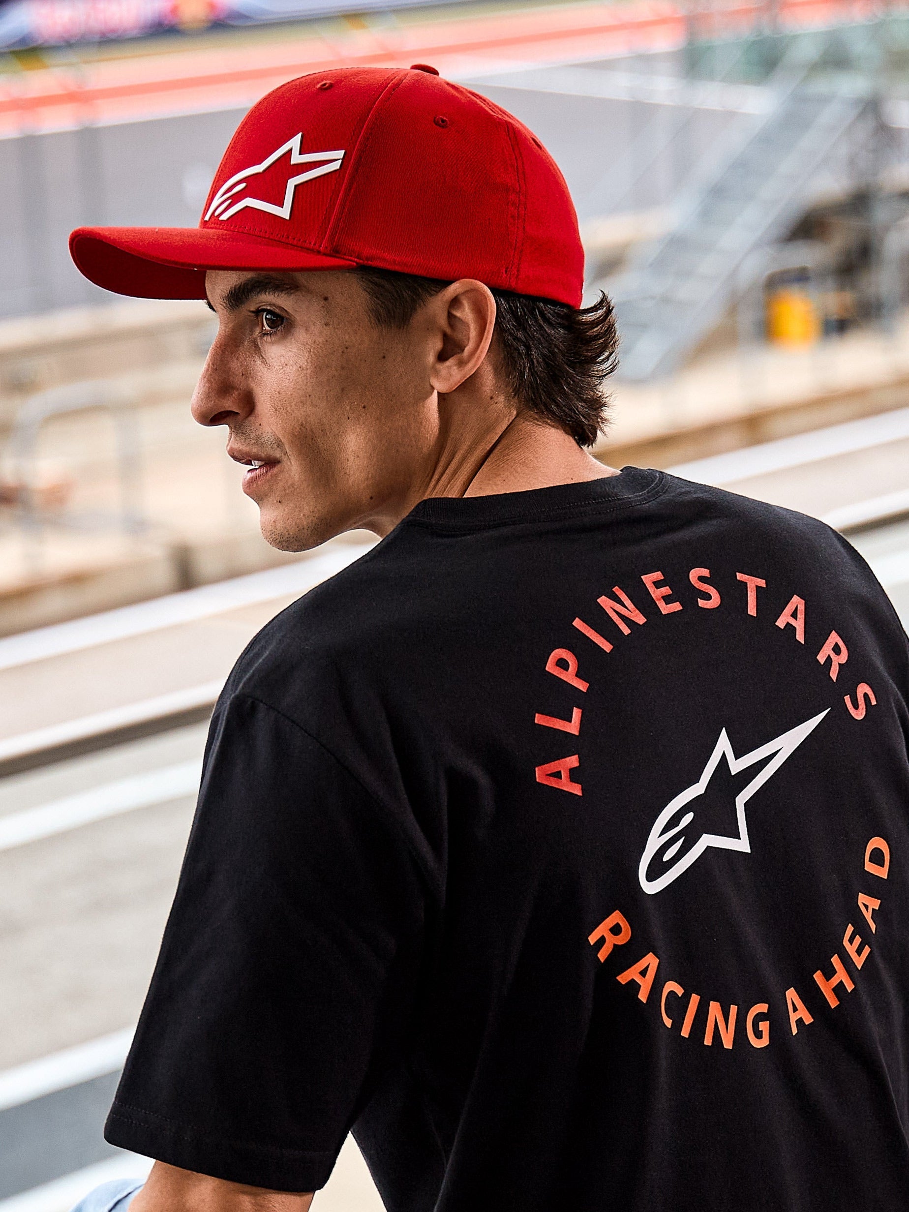 Alpinestars Corp Shift Multi Hat, boné vermelho de pala curva com logótipo Astars 3D branco, usado com uma t-shirt Alpinestars preta com o grafismo 'Racing Ahead', num cenário de lifestyle de desportos motorizados num circuito de corridas