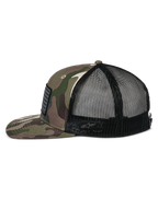 Boné Trucker Alpinestars Flag Flat, camuflado verde e preto, aba plana e coroa estruturada, frente em sarja camuflada com malha preta na parte traseira, vista lateral mostrando remendo de bandeira merrow e logótipo bordado, streetwear casual inspirado no desporto motorizado