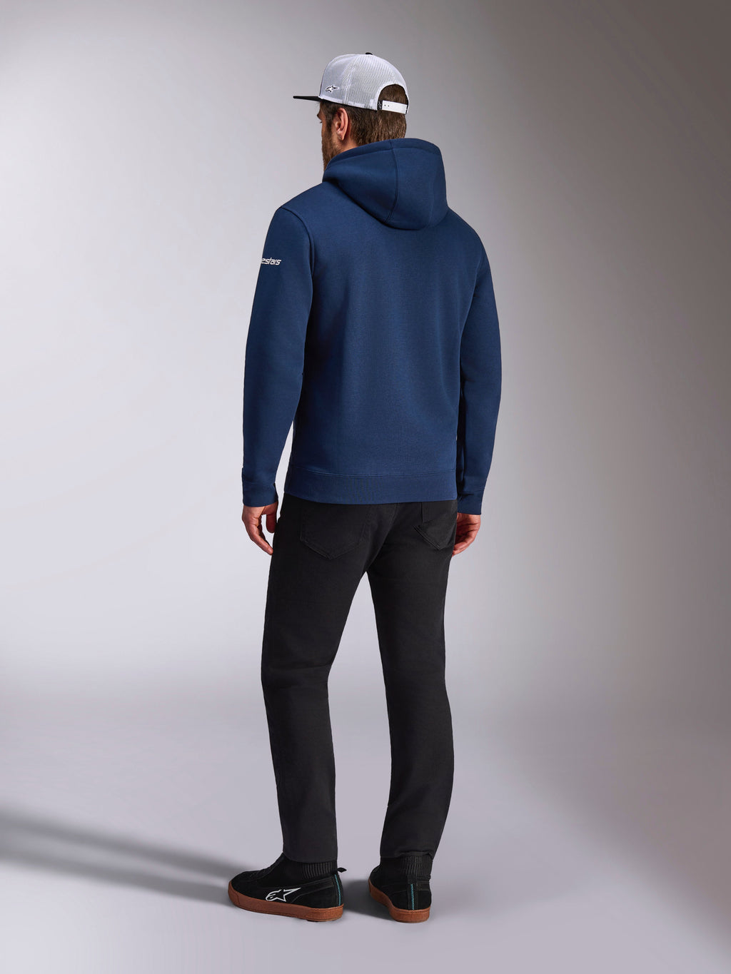 Alpinestars Frontal Hoodie, vista traseira de um hoodie de lã polar azul-marinho, logótipo branco na manga, combinado com calças pretas e um boné trucker de rede branco, streetwear casual inspirado no desporto motorizado