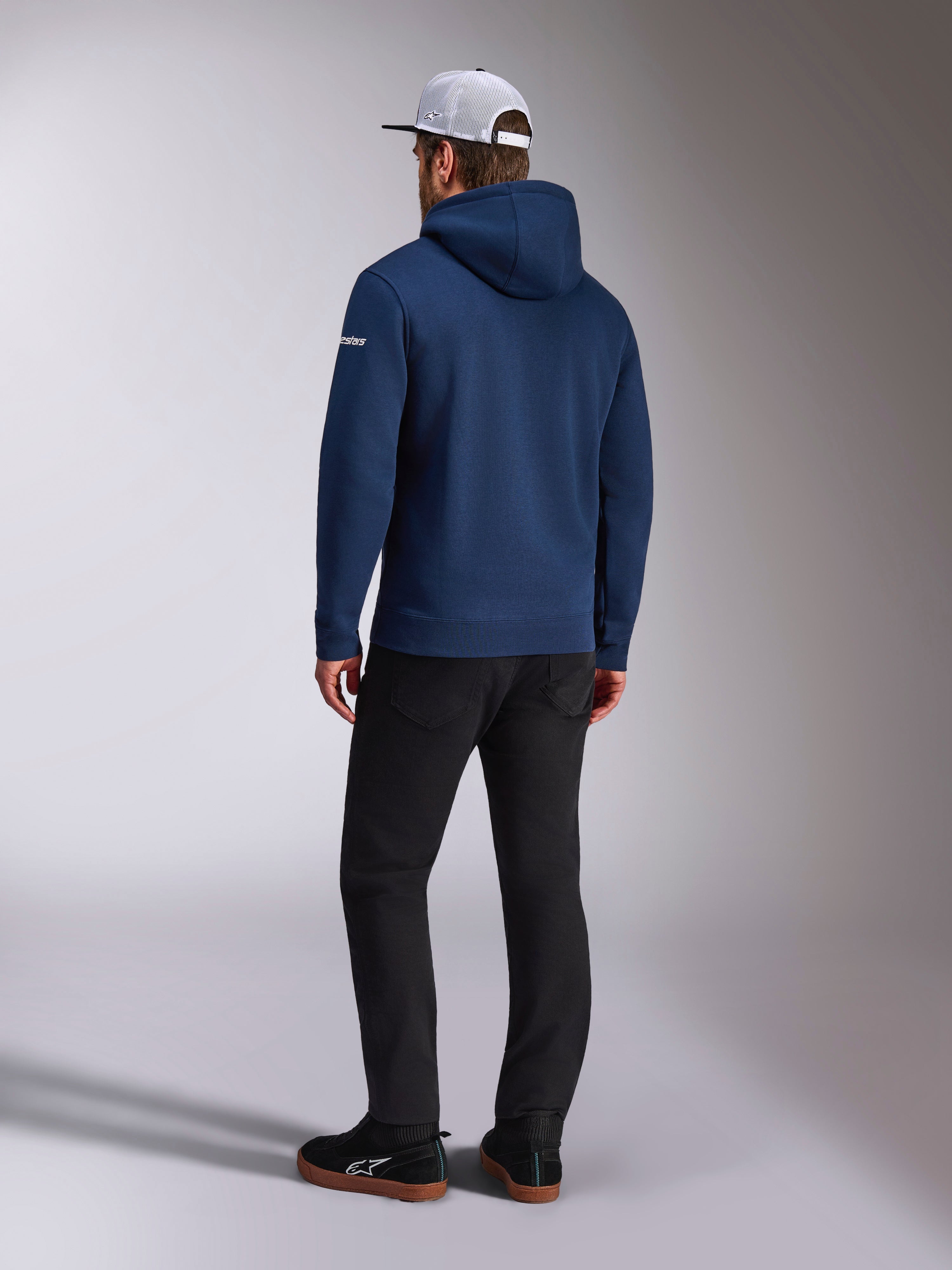Alpinestars Frontal Hoodie, vista traseira de um hoodie de lã polar azul-marinho, logótipo branco na manga, combinado com calças pretas e um boné trucker de rede branco, streetwear casual inspirado no desporto motorizado