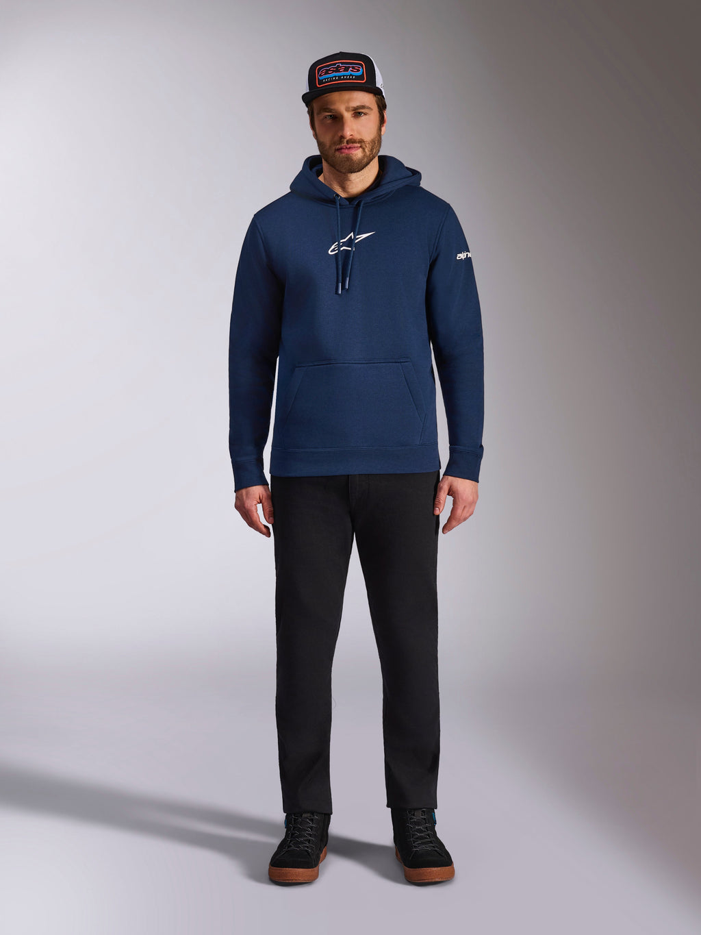 Modelo masculino a usar o Hoodie Frontal da Alpinestars em azul-marinho, sweatshirt de lã polar com logótipo Astars branco no peito e logótipo da marca na manga, com bolso tipo canguru e capuz com cordão, combinado com calças pretas e boné da marca, streetwear casual de desporto motorizado