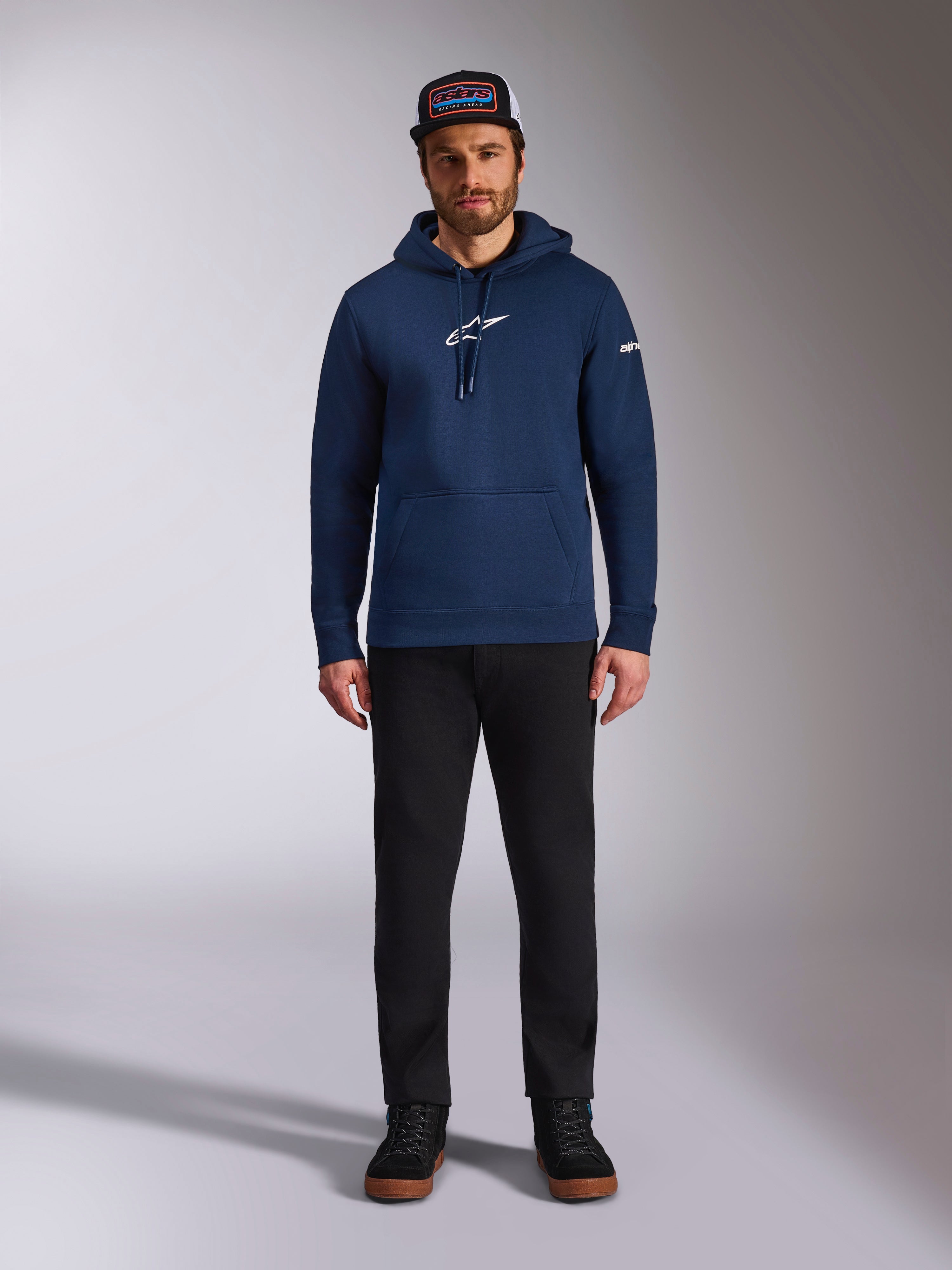 Modelo masculino a usar o Hoodie Frontal da Alpinestars em azul-marinho, sweatshirt de lã polar com logótipo Astars branco no peito e logótipo da marca na manga, com bolso tipo canguru e capuz com cordão, combinado com calças pretas e boné da marca, streetwear casual de desporto motorizado