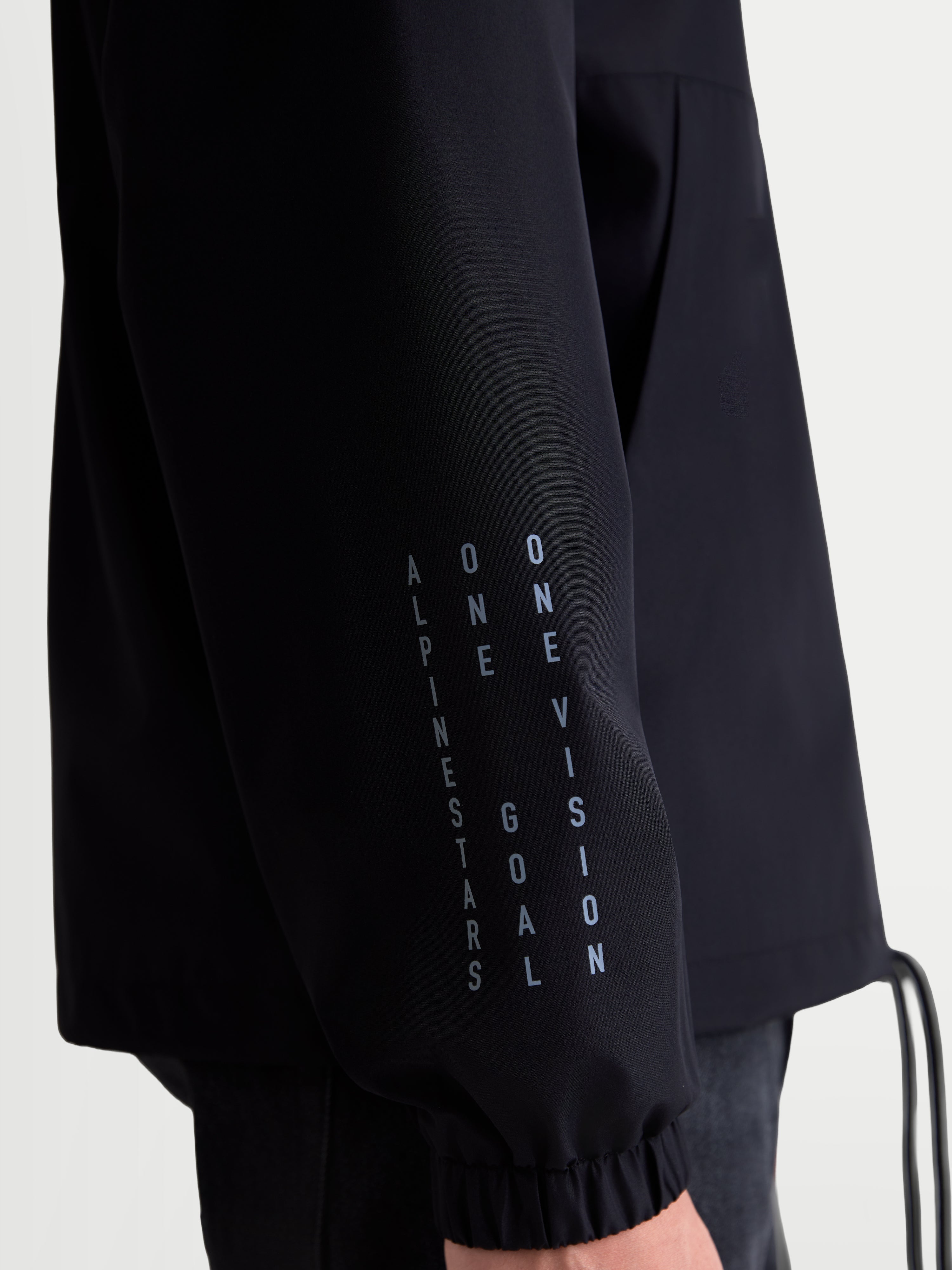 Alpinestars casaco corta-vento Speeder Resolve, casaco de sportswear leve em preto, vista de pormenor da manga com marca vertical Alpinestars, One Goal e One Vision, punho elástico, construção em poliéster para uso urbano.