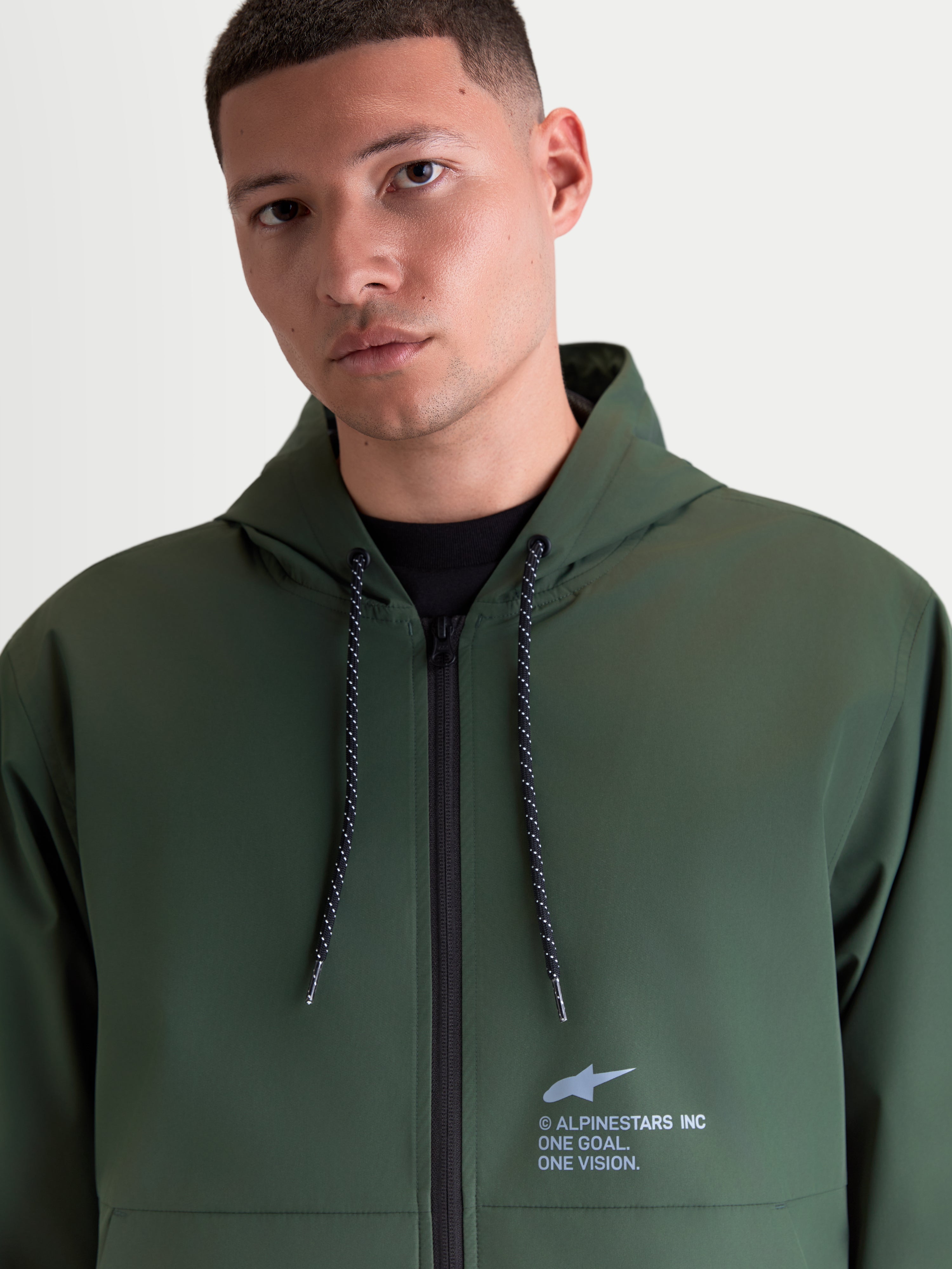 Alpinestars Speeder Resolve Corta-Vento, casaco leve com capuz para homem, verde, com cordões ajustáveis salpicados e marca Alpinestars no peito, casual sportswear, vestido por um modelo masculino.