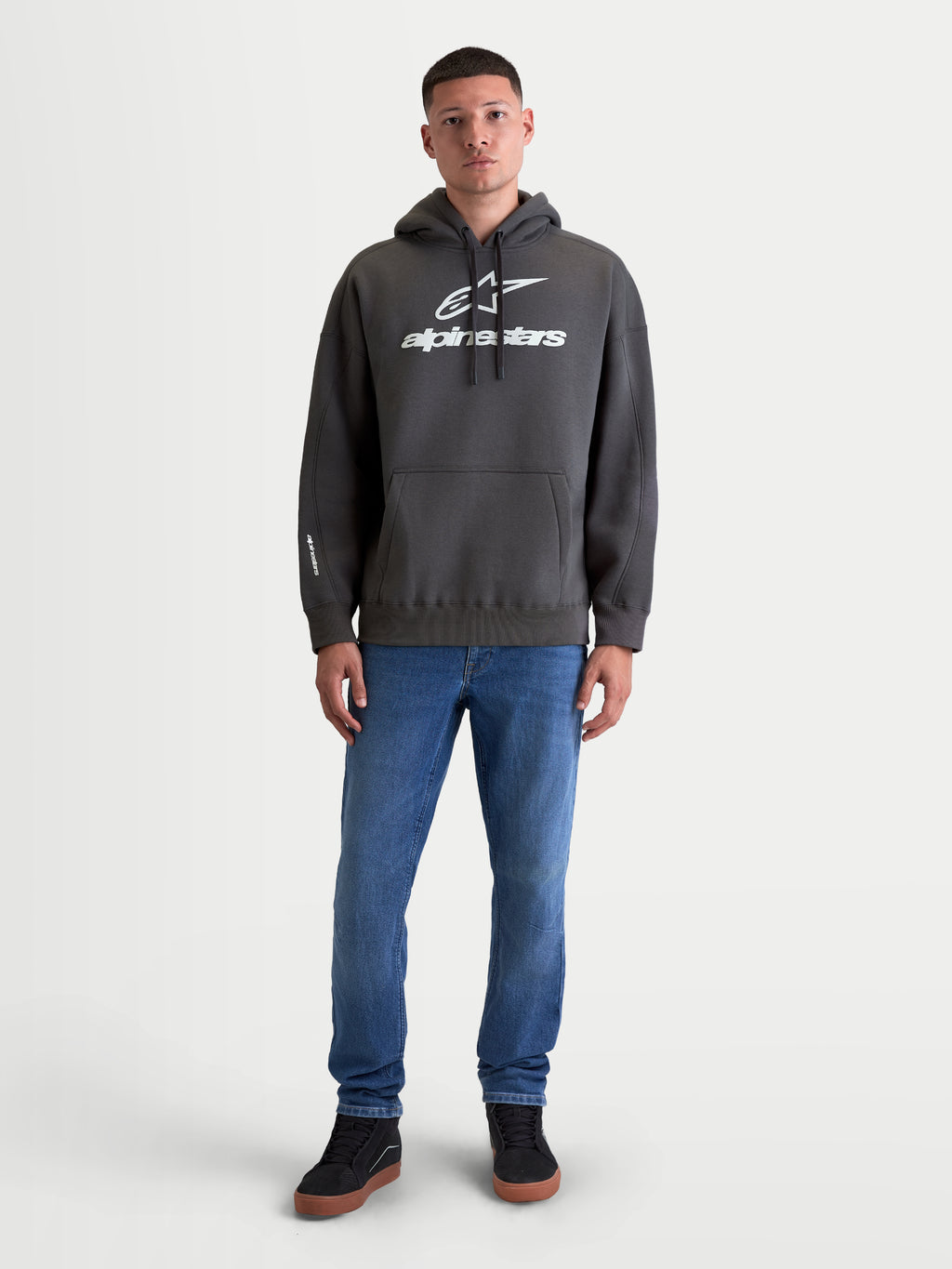 Alpinestars Camisola Com Capuz Prelude Oversized, hoodie de fleece unissexo em cinzento-escuro, com corte oversized descontraído, bolso canguru e logótipos em relevo brancos no peito e na manga, apresentada em modelo para um visual casual de dia-a-dia.