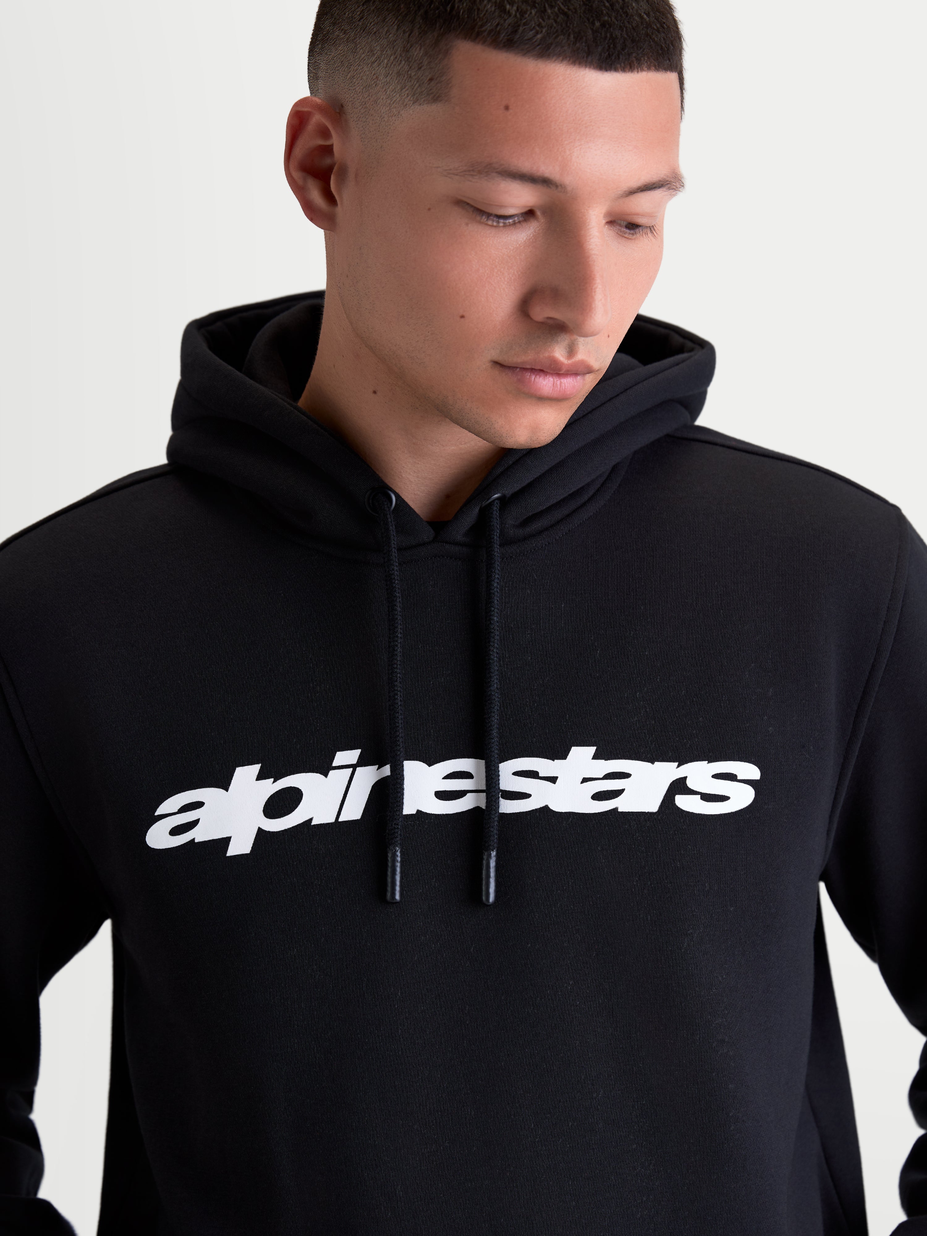 Alpinestars Camisola Com Capuz Race Wordmark vestida por um modelo, hoodie de estilo sportswear preta e branca, com logótipo wordmark frontal, capuz forrado e pontas do cordão mergulhadas, construção em algodão e poliéster reciclado.