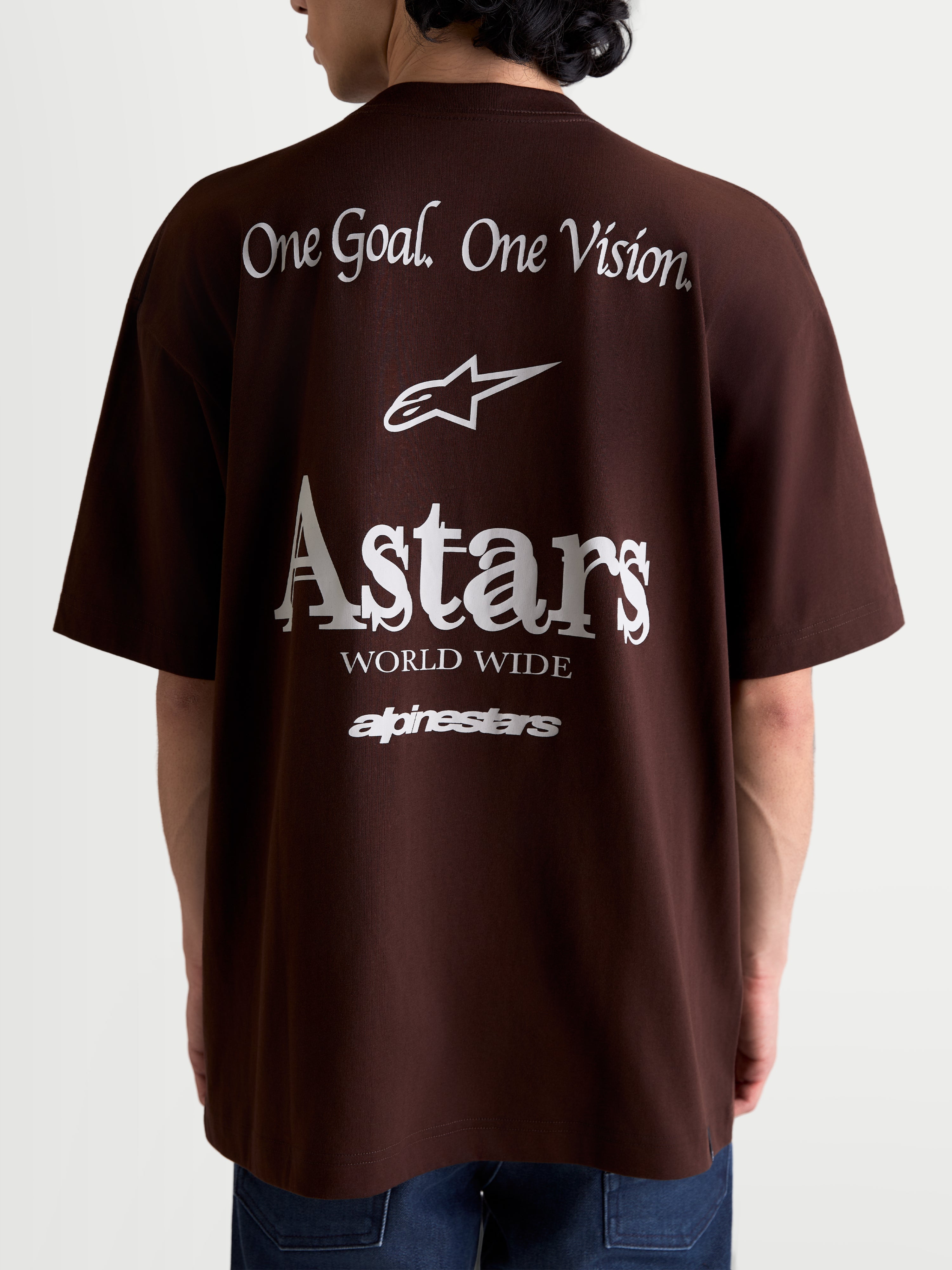 Alpinestars T-Shirt Post Oversized CSF, t-shirt de manga curta castanha, vista traseira num modelo com gráficos brancos 'One Goal. One Vision.' e 'Astars World Wide', corte relaxado em mistura de algodão e poliéster, sportswear