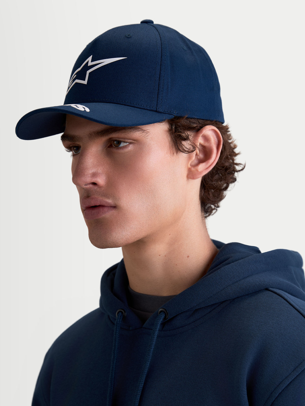 Alpinestars Arclight Hat, boné sportswear azul marinho para homem e mulher, com aba curva, logótipo Astars em relevo branco e copa estruturada. Vista de perfil em modelo mostrando o ajuste clássico inspirado no motorsport e o tecido de sarja de algodão resistente.