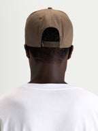 Alpinestars Boné Acme Snapback, boné casual castanho walnut usado virado para trás num modelo, mostrando o fecho snapback ajustável e a etiqueta traseira com o logótipo