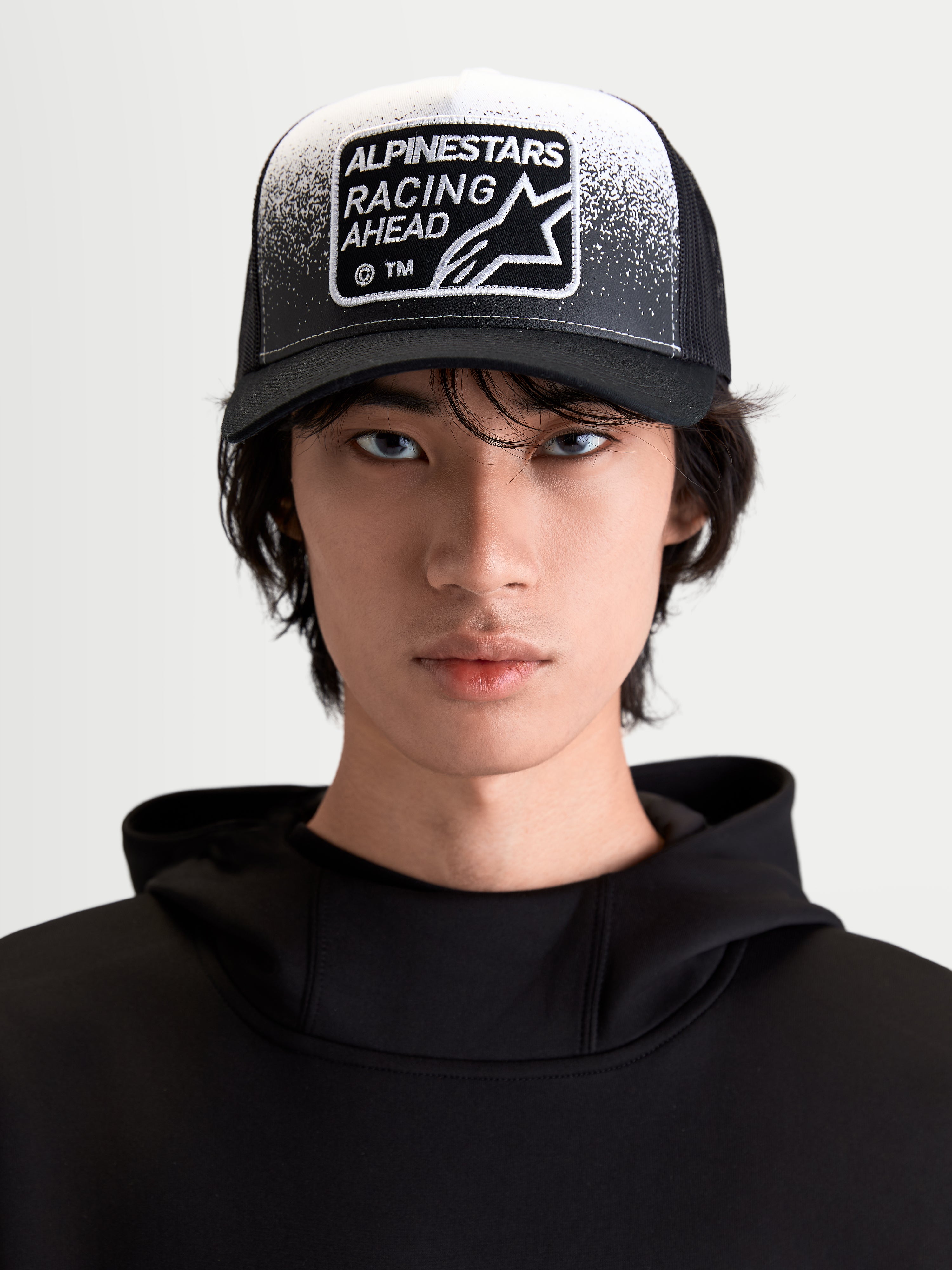 Alpinestars boné trucker Gravel, boné sportswear lifestyle, preto e branco, vista frontal no modelo com pala curva, malha traseira respirável e patch tecido Racing Ahead com detalhe de textura de cascalho.