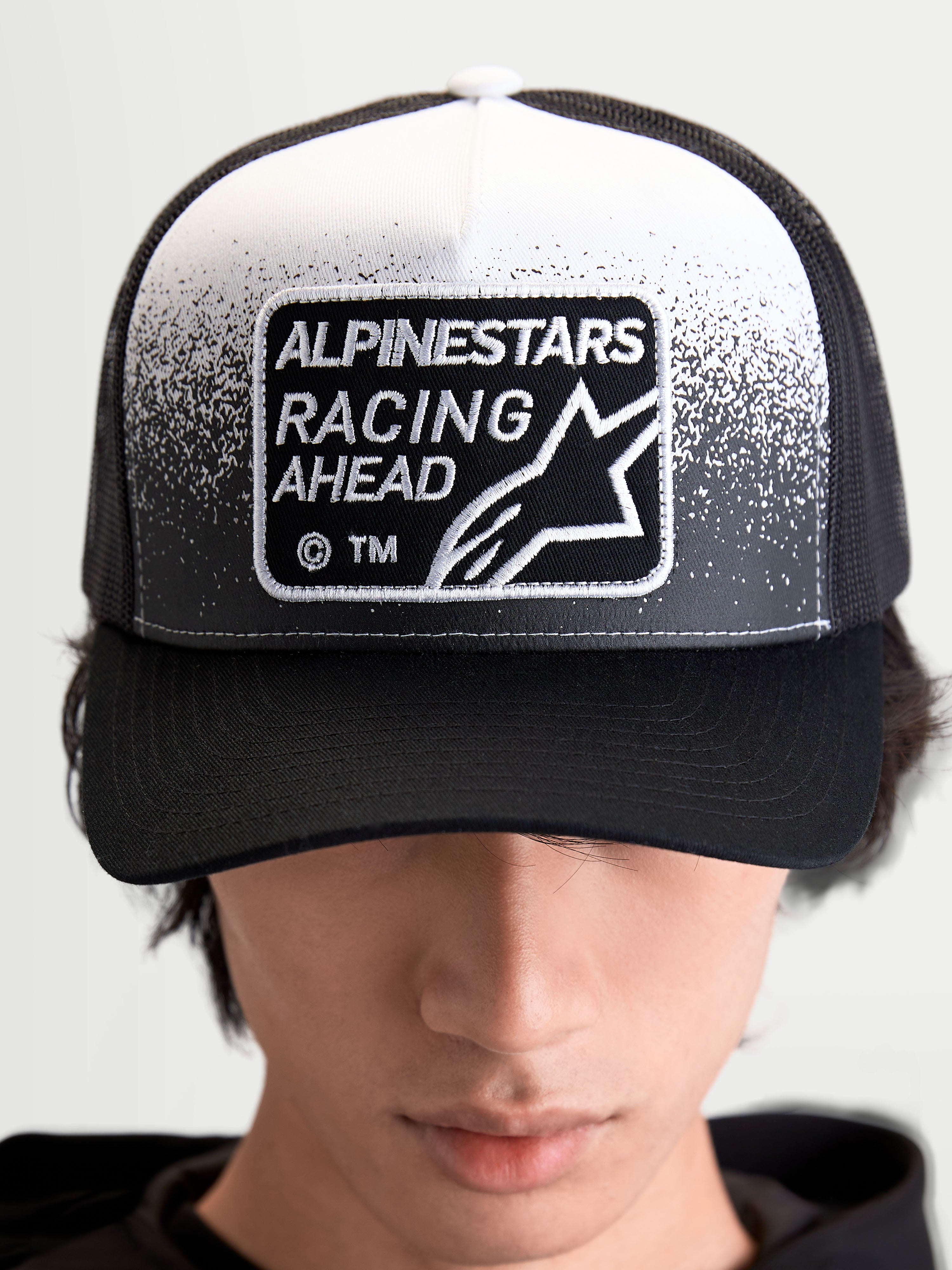 Alpinestars Boné Trucker Gravel, boné unissexo de estilo sportswear, preto e branco, com patch tecido Alpinestars Racing Ahead e detalhe de textura de gravilha, copa estruturada e aba curva, vista frontal num modelo.