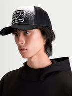 Alpinestars Boné Trucker Gravel, boné sportswear casual em modelo, preto e branco, com copa estruturada, pala curva, malha traseira respirável e patch tecido frontal com textura de cascalho e marca Alpinestars Racing Ahead.