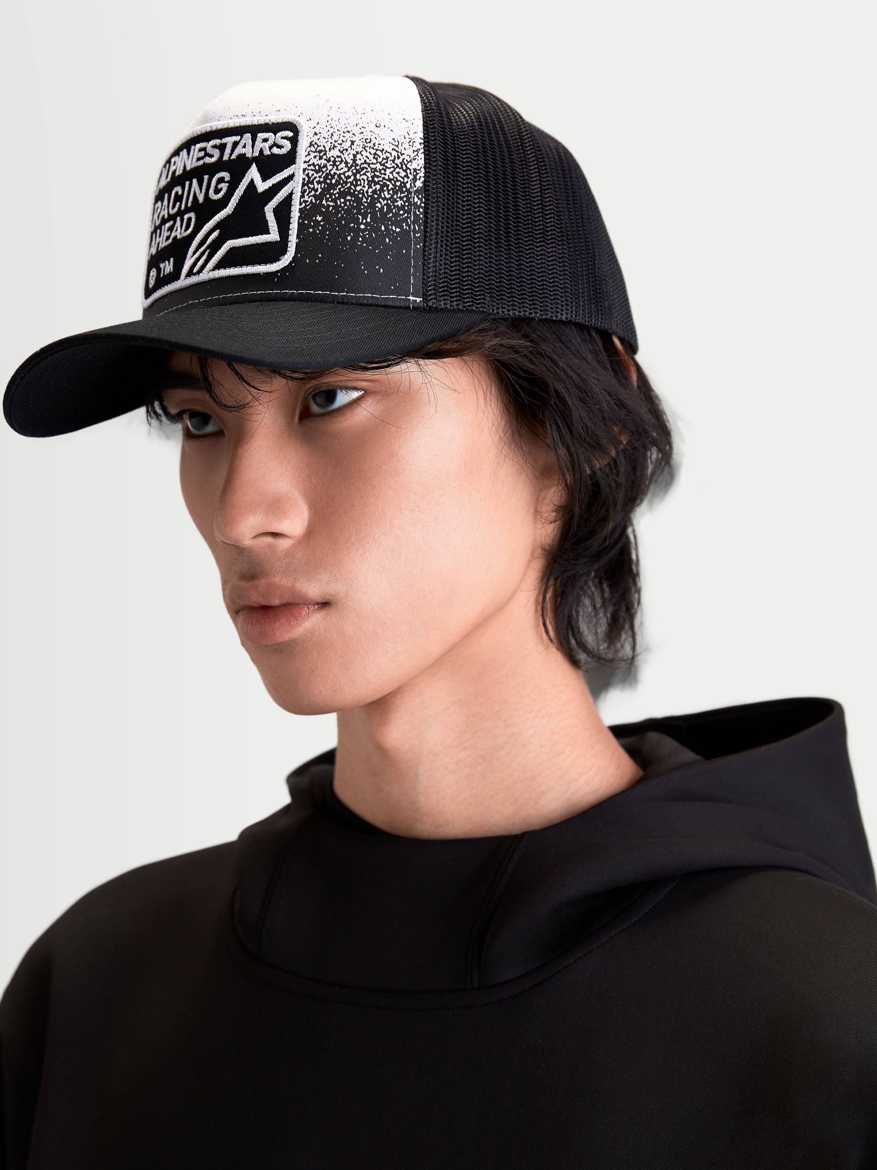 Alpinestars Boné Trucker Gravel, boné sportswear casual em modelo, preto e branco, com copa estruturada, pala curva, malha traseira respirável e patch tecido frontal com textura de cascalho e marca Alpinestars Racing Ahead.