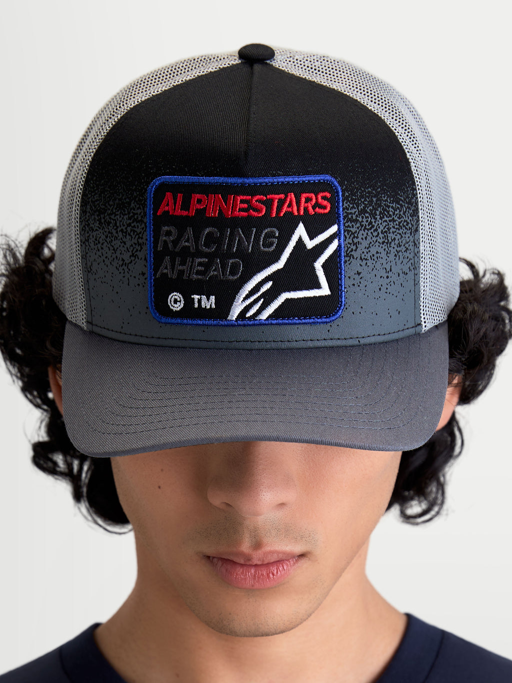 Alpinestars Boné Trucker Gravel, boné de malha de sportswear, cinzento escuro e preto, com pala curva e patch tecido frontal com a inscrição 'Racing Ahead' e o logótipo Astars, vista frontal num modelo.