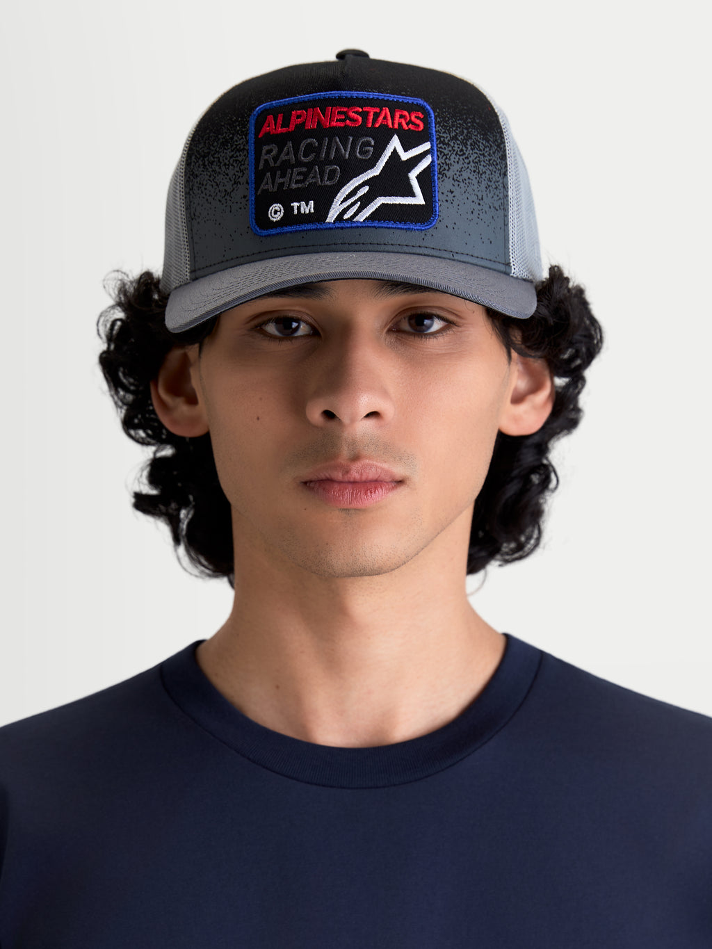 Alpinestars Boné Trucker Gravel, boné de sportswear unissexo em cinzento escuro e preto, visto num modelo. Apresenta copa estruturada, aba curva, malha respirável na parte traseira e um patch frontal tecido com a marca Alpinestars Racing Ahead e detalhe de textura de gravilha.