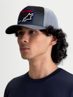 Alpinestars boné trucker Gravel, acessório casual num modelo masculino, cinzento escuro e preto com malha cinzenta clara atrás, copa estruturada, pala curva e patch frontal tecido com o logótipo Alpinestars e texto vermelho, estilo sportswear inspirado na competição.
