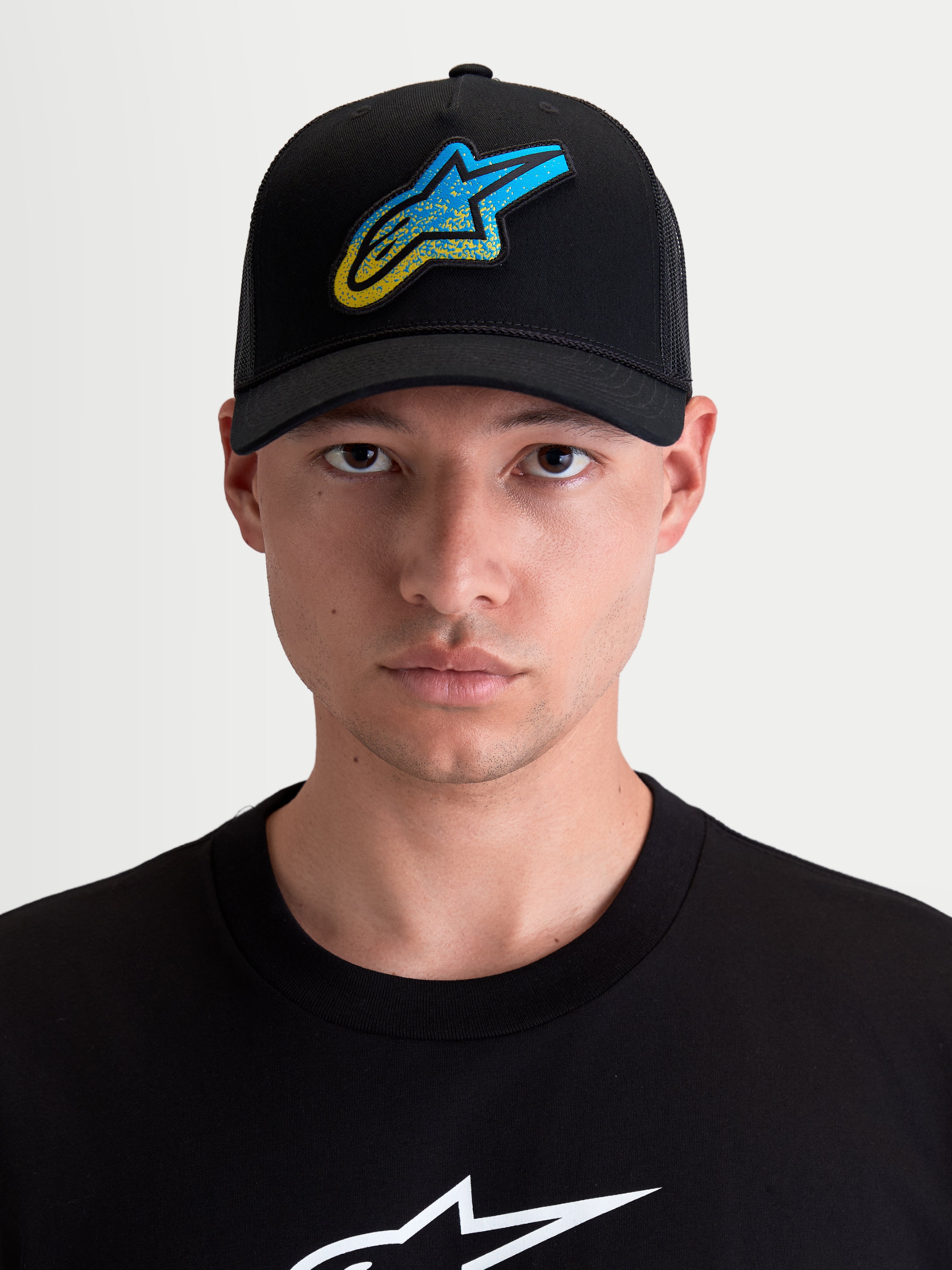 Alpinestars boné trucker Meteorite, boné de sportswear unissexo com pala curva, preto com logótipo Astars azul e amarelo com textura de cascalho, frente em sarja e malha traseira em poliéster, modelo a usar o boné em vista frontal.