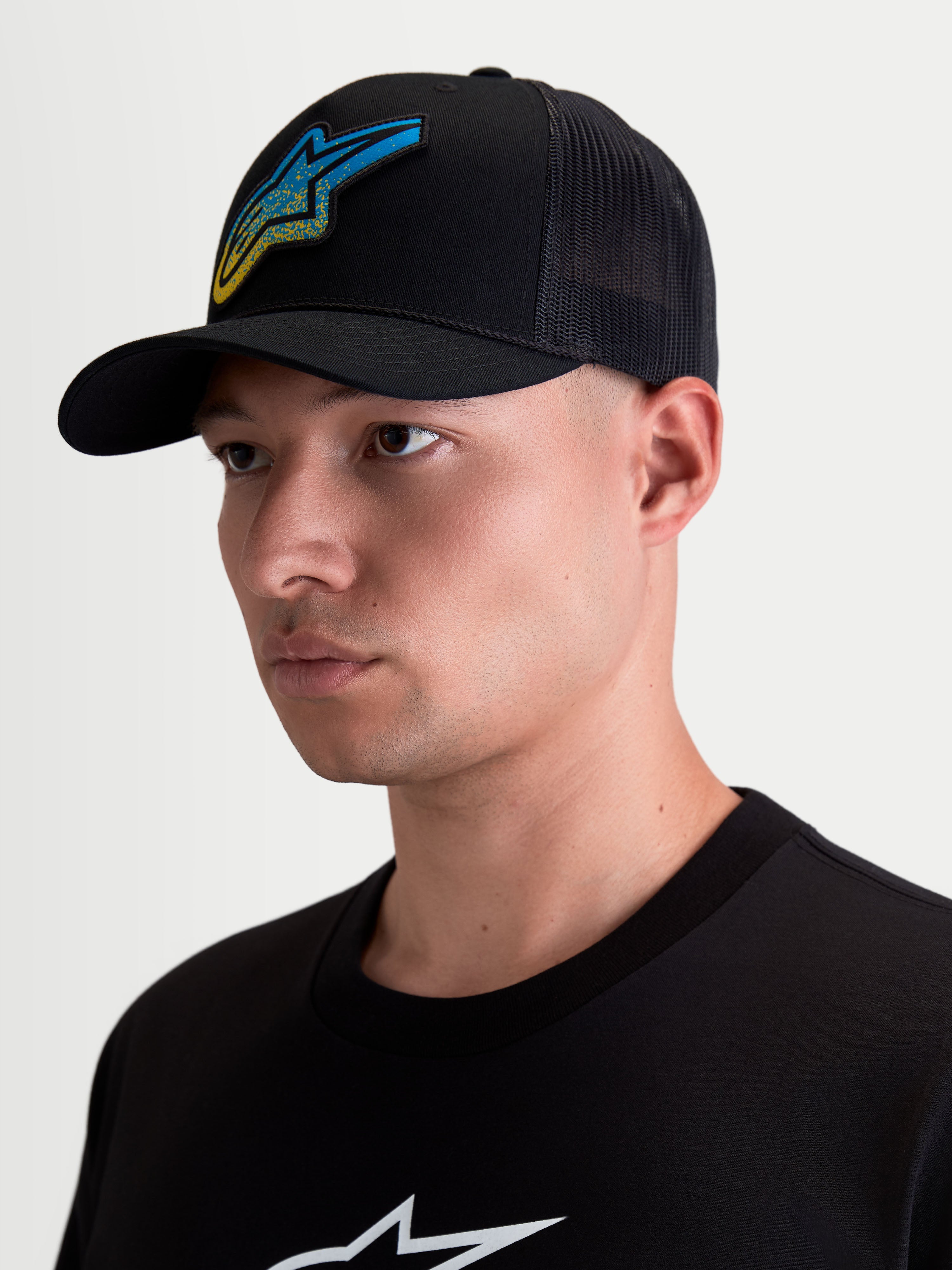 Boné Trucker Meteorite Alpinestars, boné de sportswear casual para homem, preto com logótipo Astars tecido em azul e amarelo, pala curva e coroa estruturada, parte traseira em malha respirável, mostrado num modelo.