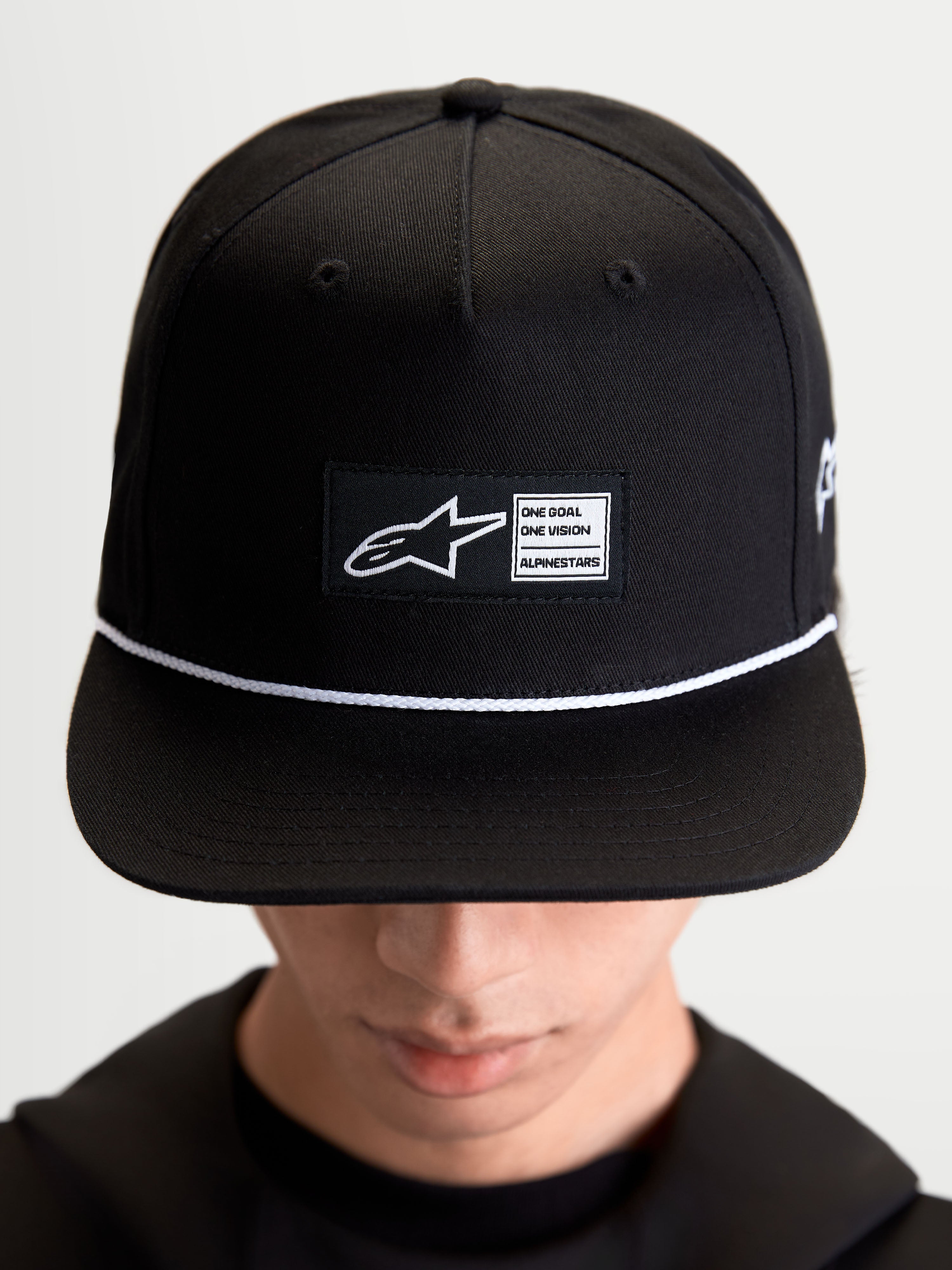Alpinestars boné Label Snapback, acessório sportswear lifestyle unissexo, preto e branco, em sarja de algodão, com copa estruturada, aba plana, detalhe de cordão branco e etiqueta tecida frontal com branding Alpinestars, mostrado no modelo.