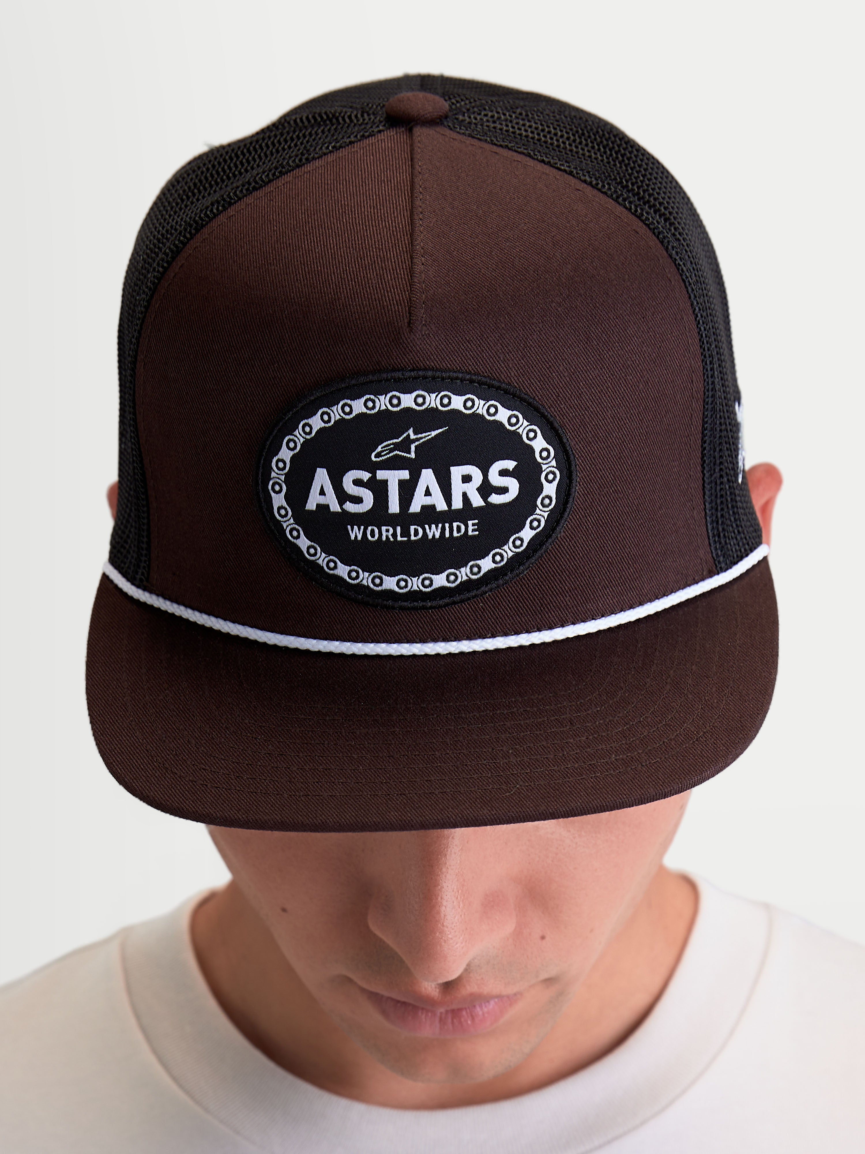 Alpinestars Boné Trucker Hotlink, boné de sportswear inspirado no motorsport mostrado num modelo, castanho e preto, com copa estruturada, aba plana com detalhe de cordão branco e patch frontal com logótipo Astars Worldwide com rebordo de corrente de bicicleta.