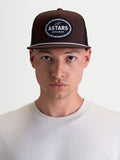 Alpinestars Boné Trucker Hotlink, boné de sportswear inspirado no motorsport, castanho e preto, copa estruturada, aba plana com detalhe de corda branca e patch frontal com logótipo Astars Worldwide, apresentado num modelo.