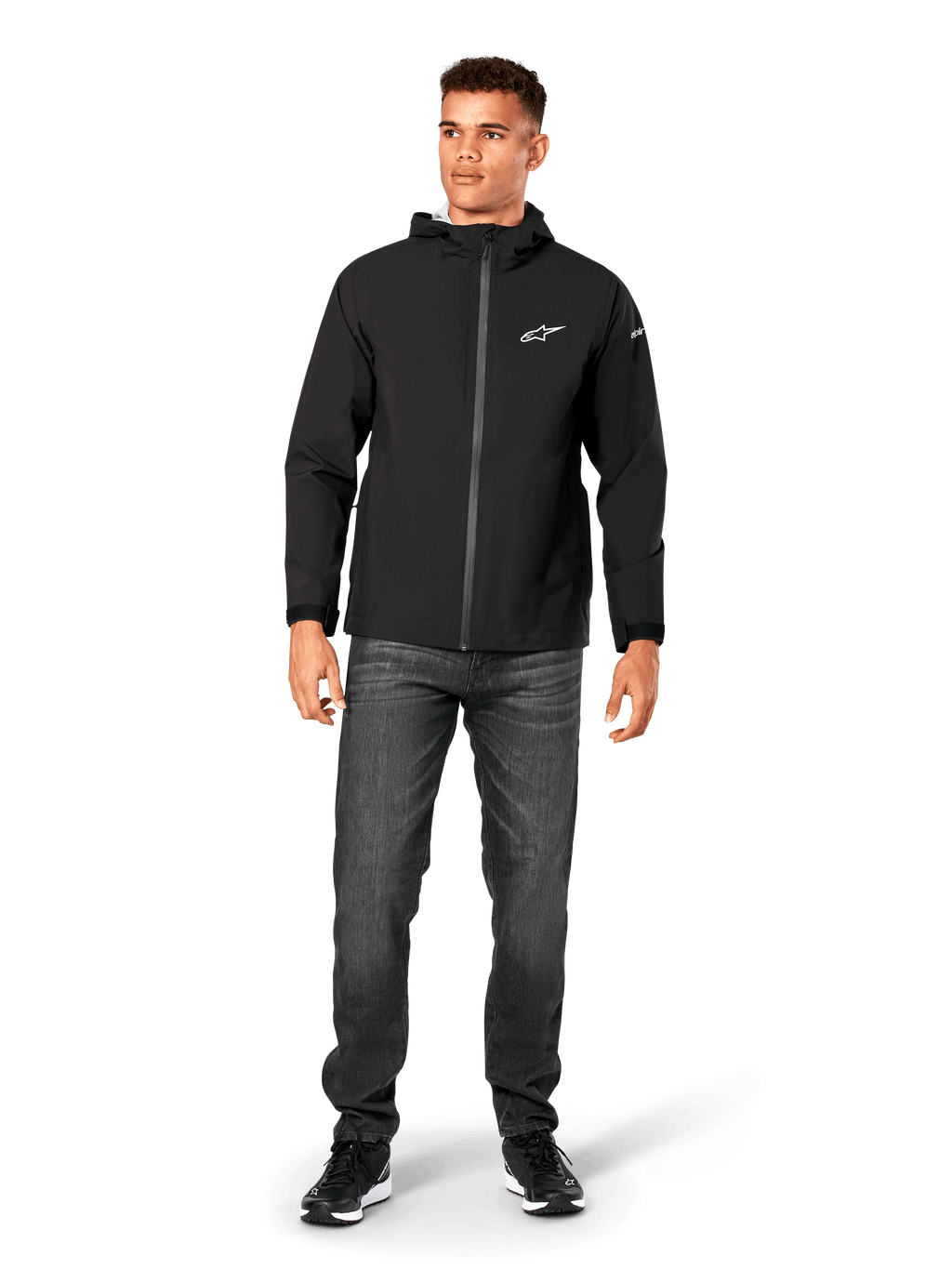 Alpinestars Kascade Rain Jacket, casaco impermeável para homem, preto, usado com calças de ganga cinzento-escuras e sapatilhas pretas, com capuz, fecho frontal revestido e logótipo Alpinestars branco no peito, streetwear casual inspirado no desporto motorizado