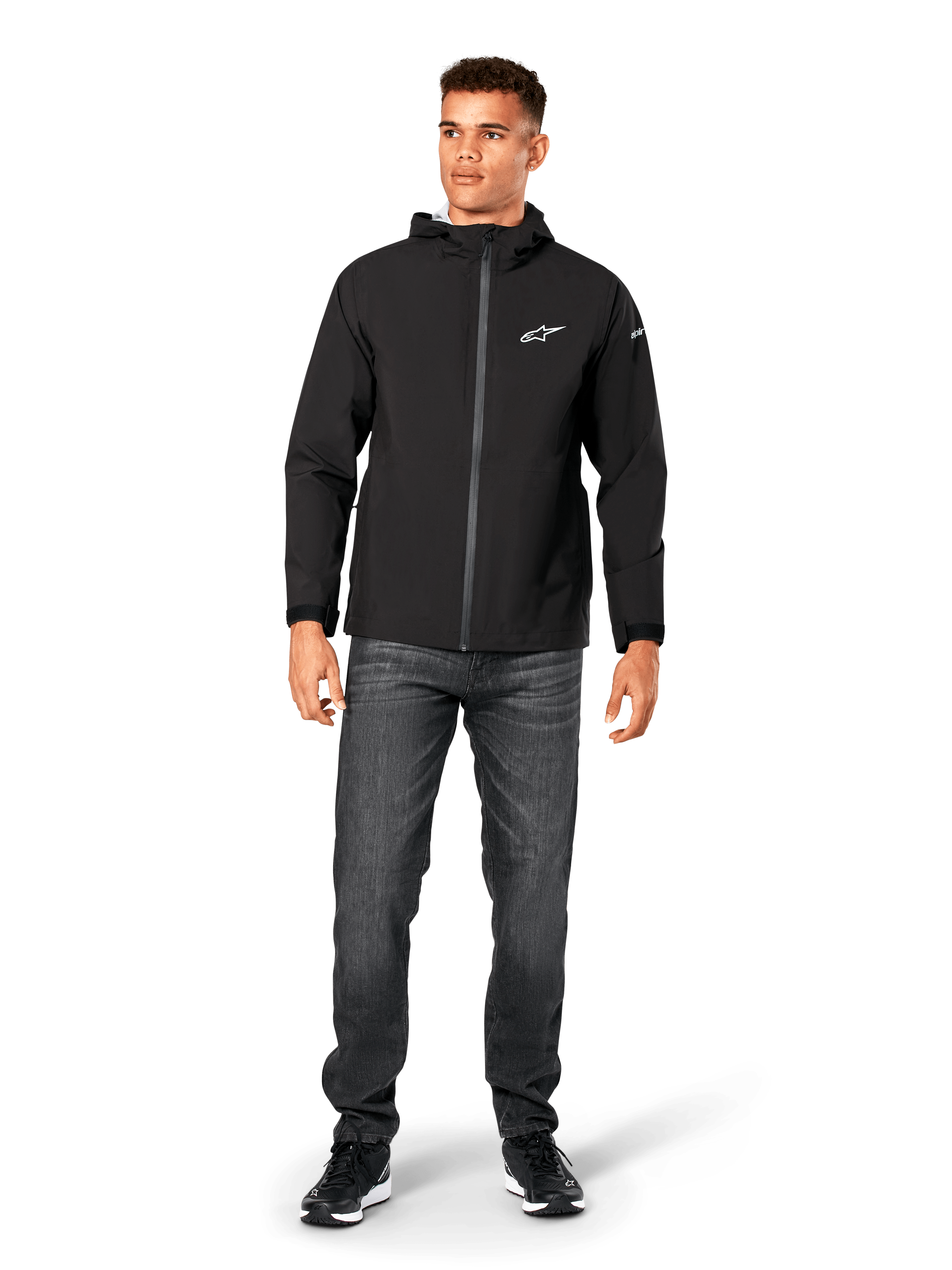 Alpinestars Kascade Rain Jacket, casaco impermeável para homem, preto, usado com calças de ganga cinzento-escuras e sapatilhas pretas, com capuz, fecho frontal revestido e logótipo Alpinestars branco no peito, streetwear casual inspirado no desporto motorizado