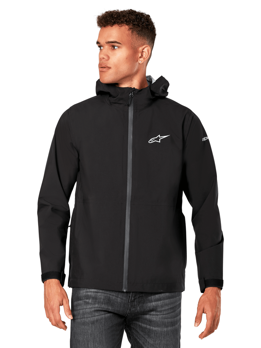 Modelo masculino a vestir o casaco impermeável Alpinestars Kascade em preto, com logótipo branco no peito e marca na manga, casaco com capuz, fecho frontal revestido e punhos com ajuste de velcro, combinado com calças de ganga escuras, num estilo streetwear casual inspirado no desporto motorizado.