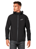 Modelo masculino a vestir o casaco impermeável Alpinestars Kascade em preto, com logótipo branco no peito e marca na manga, casaco com capuz, fecho frontal revestido e punhos com ajuste de velcro, combinado com calças de ganga escuras, num estilo streetwear casual inspirado no desporto motorizado.