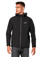 Modelo masculino a vestir o casaco impermeável Alpinestars Kascade em preto, com logótipo branco no peito e marca na manga, casaco com capuz, fecho frontal revestido e punhos com ajuste de velcro, combinado com calças de ganga escuras, num estilo streetwear casual inspirado no desporto motorizado.