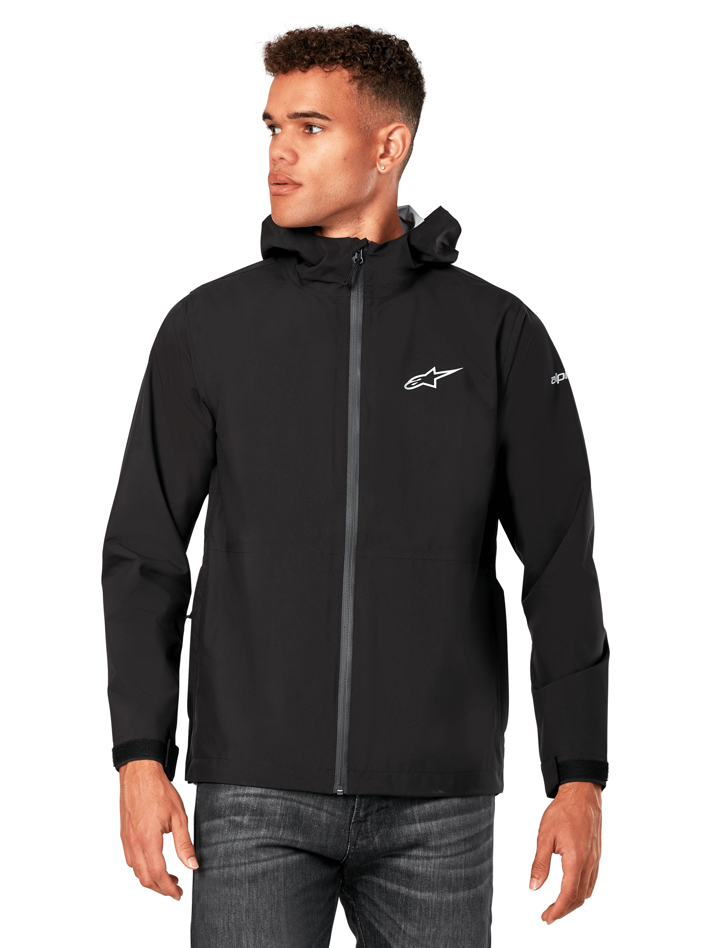 Modelo masculino a vestir o casaco impermeável Alpinestars Kascade em preto, com logótipo branco no peito e marca na manga, casaco com capuz, fecho frontal revestido e punhos com ajuste de velcro, combinado com calças de ganga escuras, num estilo streetwear casual inspirado no desporto motorizado.
