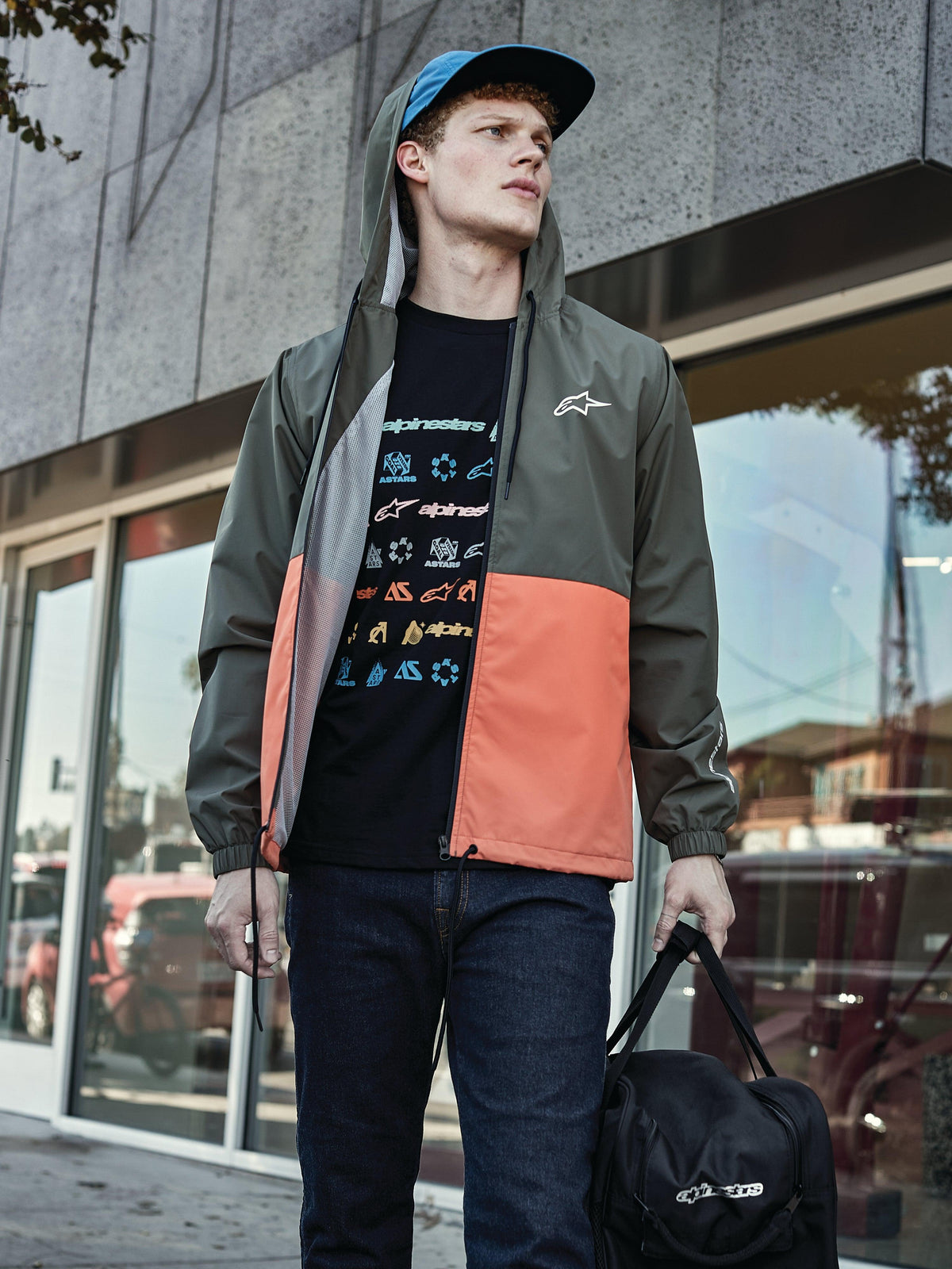 Homem a usar o casaco corta-vento Alpinestars Speeder em Military Orange, com design de blocos de cor em verde tropa e laranja, capuz com forro de malha, t-shirt preta com logótipo gráfico, calças de ganga azuis e saco de desporto preto Alpinestars, visual casual lifestyle
