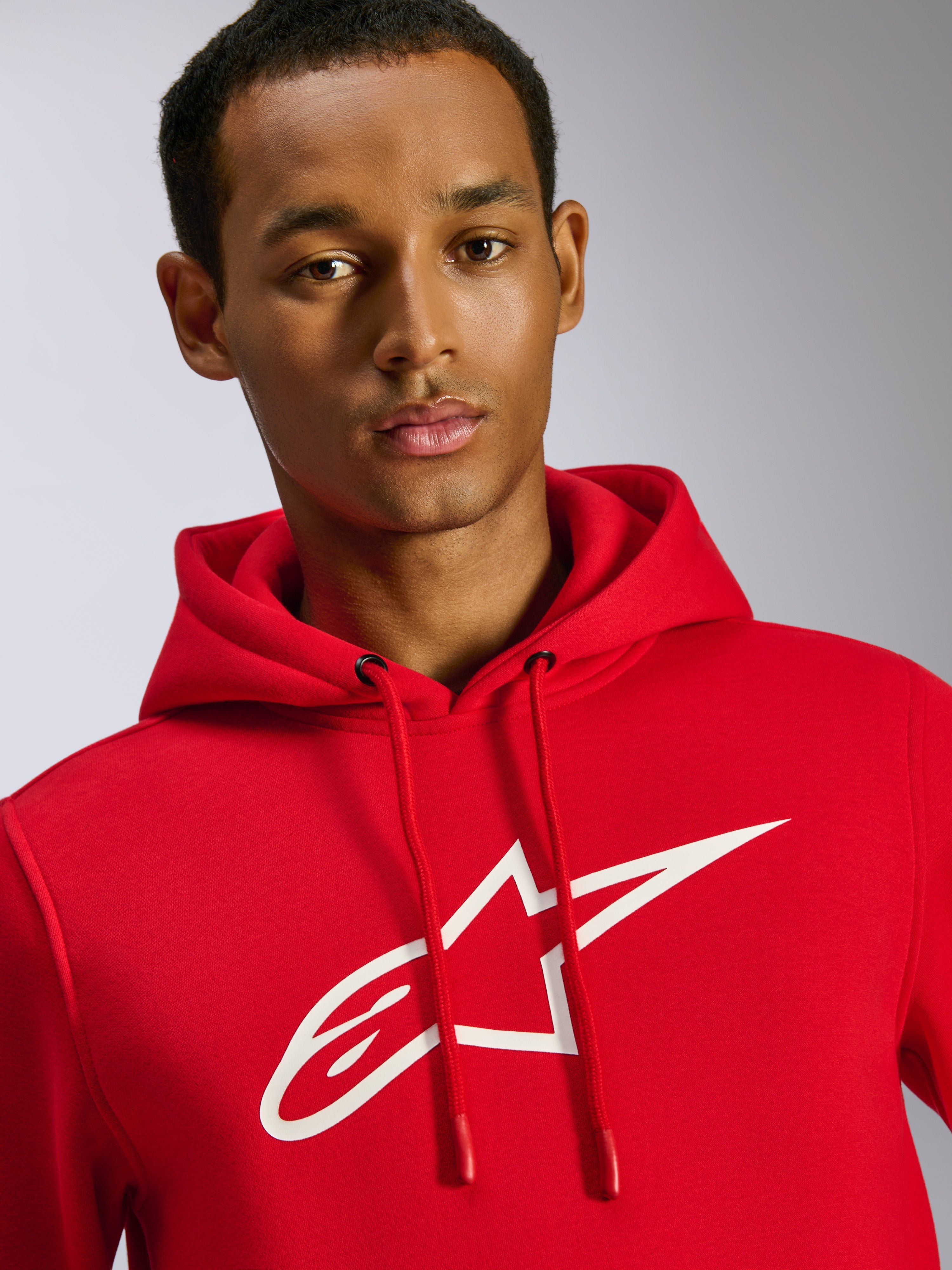 Alpinestars Ageless V3 Hoodie, camisola com capuz vermelha, com um logótipo Alpinestars grande em branco no peito, capuz com cordão e pontas mergulhadas, streetwear casual inspirado no desporto motorizado, vestido por um modelo masculino