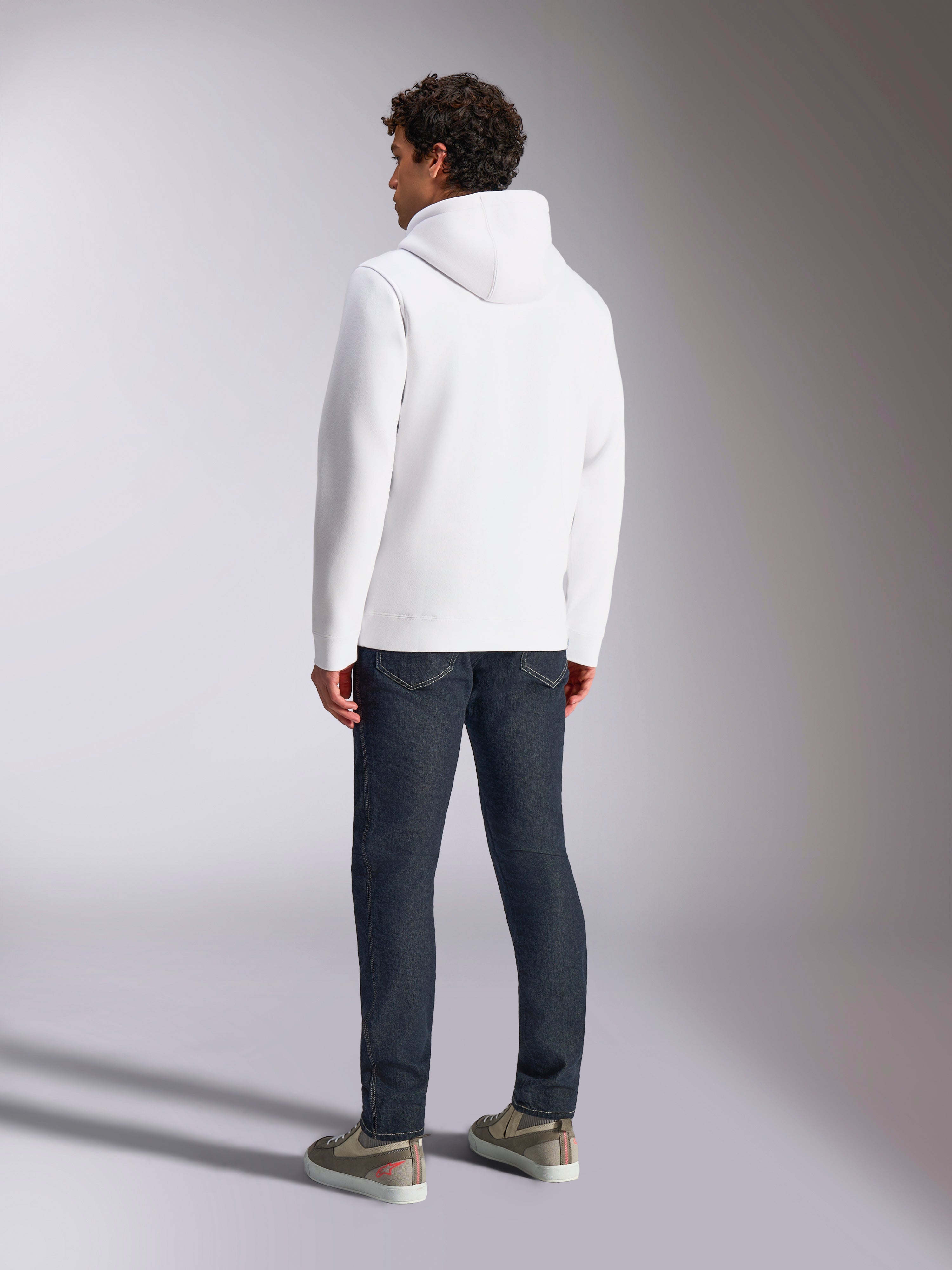 Alpinestars Blaze V3 Hoodie, vista traseira de modelo masculino a usar uma camisola com capuz branca, roupa desportiva casual combinada com calças de ganga azul-escuras e ténis cinzentos, design moderno inspirado no desporto motorizado